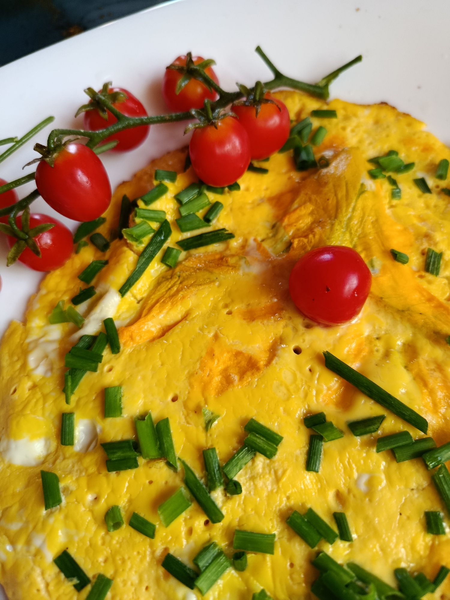 The image is a close-up, high-angle shot of an omelet on a white plate, garnished with chopped chives and cherry tomatoes on the vine. The omelet is golden yellow with visible fluffy textures and some slightly browned edges. Several small, bright red cherry tomatoes are attached to green stems, with some scattered individually on the omelet and others clustered to the left. Finely chopped green chives are sprinkled liberally over the surface of the omelet. The background is blurred but suggests a dark, possibly wooden surface. There are no people or actions depicted. The image provides no information about the city of Верея, Russia.