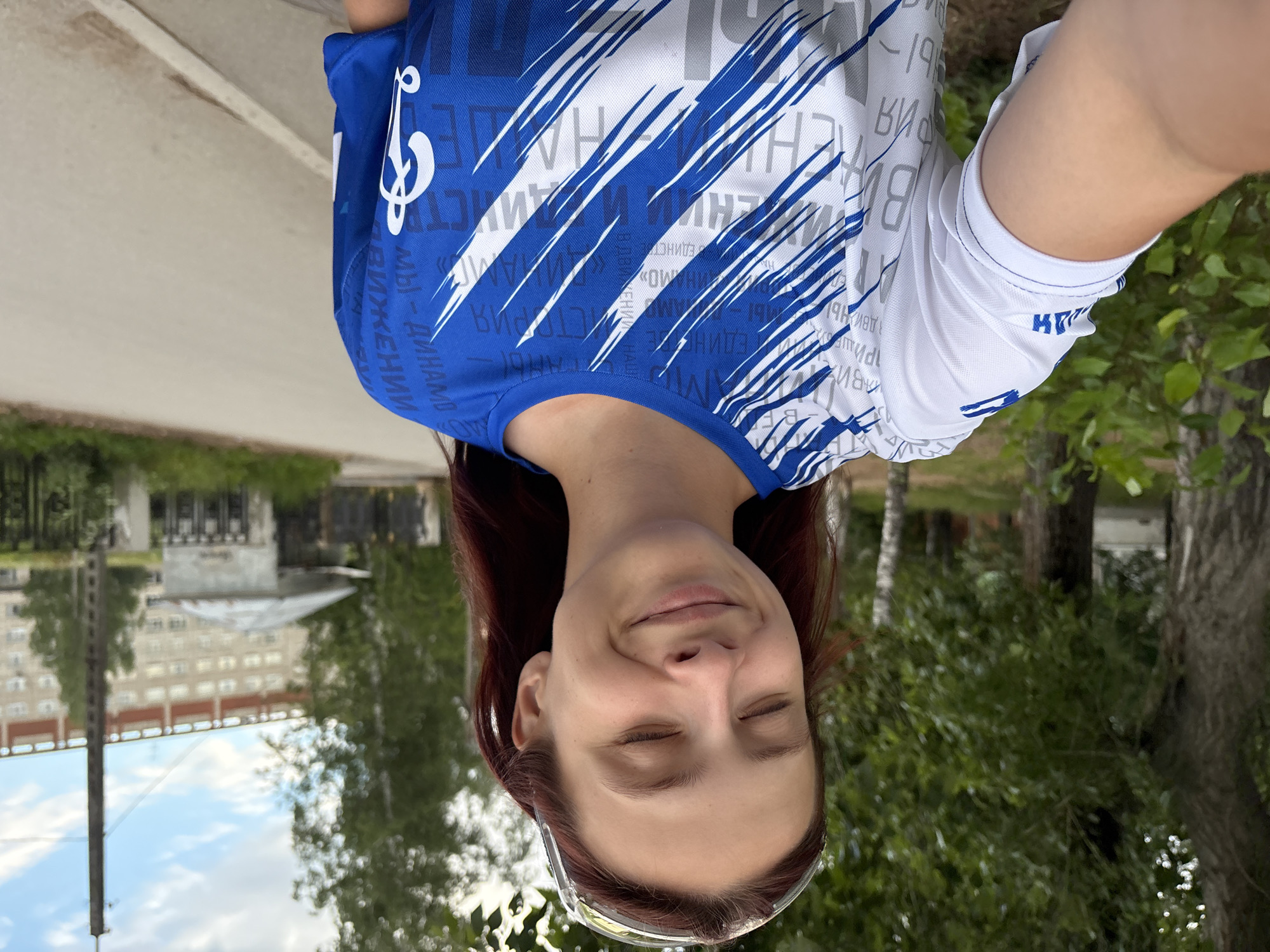 This is a portrait of a person taken from a selfie perspective. The person, a woman with reddish-brown hair, is looking at the camera with her eyes closed and a slight smile, suggesting a relaxed or happy emotion. She is wearing a blue and white t-shirt with a graphic design and text. The text on her shirt is in Russian and appears to include words like "ЕДИНСТВО" (unity) and "ДВИЖЕНИЕ" (movement). She is also wearing clear protective eyewear.
The background shows a scene outdoors, with greenery from trees and foliage visible. In the distance, there are buildings that appear to be apartment blocks, and a cloudy sky. The image is oriented vertically, with the person's head at the bottom. Based on the lighting and the greenery, it appears to be daytime, likely late morning or afternoon. The setting suggests a residential area or park in a town or city. The visible text and the style of the buildings might indicate a location in Russia, specifically Chaikovsky, Russia, as suggested by the provided location context.