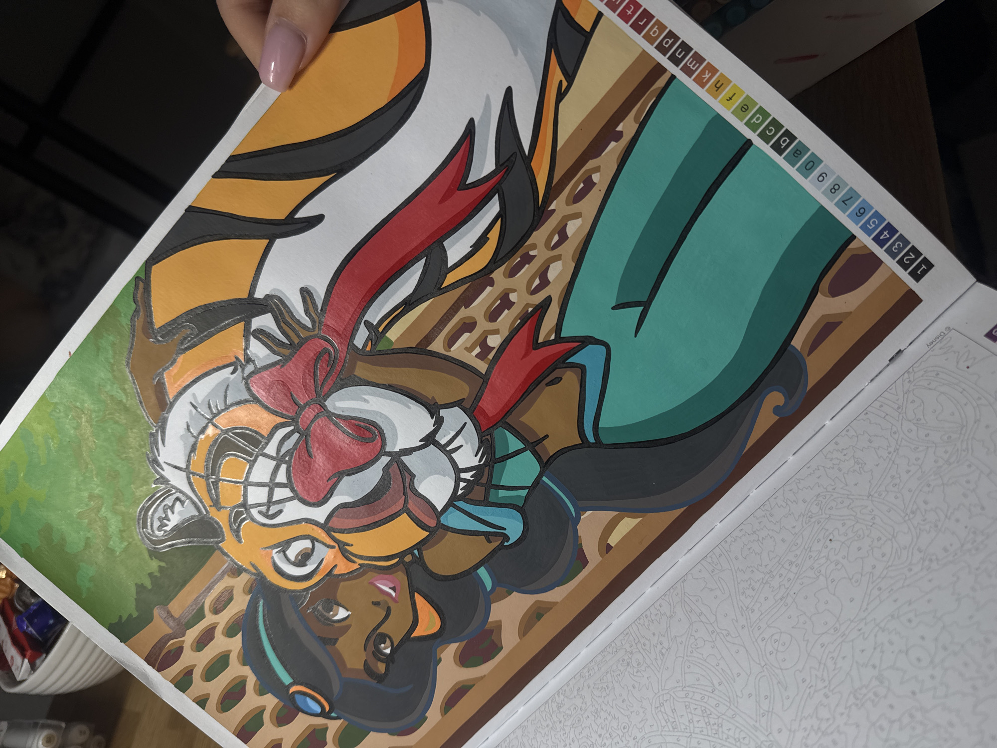 This media file shows a close-up of an open paint-by-numbers book. The foreground features a partially completed illustration of a character resembling Princess Jasmine from Disney's Aladdin, dressed as Rajah the tiger. The character has dark skin, black hair, and is wearing a tiger-striped headdress with a red bow, along with a teal outfit. The illustration is in progress, with most of the colors filled in, but the surrounding page shows the faint outline of the next section, also numbered for painting.
To the right of the illustration, along the edge of the page, there is a color key with a sequence of numbers from 1 to 9, followed by letters from 'a' to 't'. These indicate the colors to be used for the corresponding numbered and lettered sections in the painting. Some of the numbers and letters are also colored to match their respective paint shades.
In the background, a hand with a light pink polished nail is visible holding the top edge of the page. The setting appears to be indoors, with the lighting suggesting it might be daytime or well-lit artificial lighting. The overall scene captures the activity of someone engaged in a paint-by-numbers project. There is also a sliver of a white basket containing various colorful items visible in the bottom left corner.