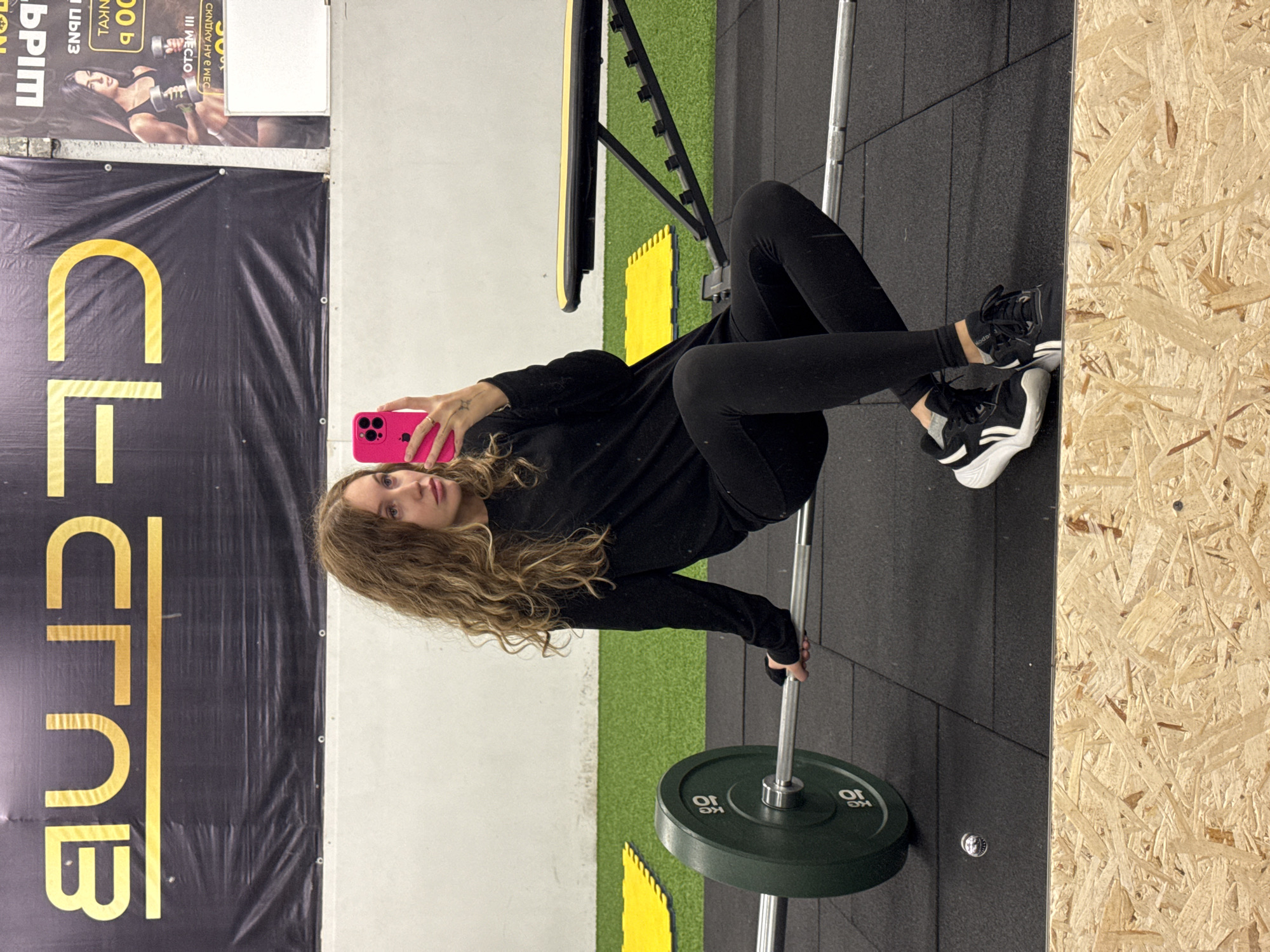 This media file depicts an indoor gym setting, rotated 90 degrees counter-clockwise. The central subject is a young woman with long, wavy, light brown hair, dressed in a black long-sleeved top, black leggings, and black and white athletic shoes. She is positioned as if performing a hip thrust exercise or resting on a weight bench, with a barbell across her hips. She is actively taking a mirror selfie, holding a bright pink smartphone towards her reflection with her right hand. Her expression is neutral and focused on her phone.
The barbell is silver with two dark green weight plates, each clearly marked "10 KG". The floor beneath her is a mix of black rubber mats and sections of green artificial turf. In the background, there is a plain white wall and a section of paneling made from light-colored chipboard or OSB.
Two banners are visible on the wall. A large black banner with gold text is on the left, displaying partial Cyrillic letters that appear to spell "FIT CLUB" when oriented correctly. Above this, a smaller poster features a muscular woman holding dumbbells. This poster contains Cyrillic text, which, when properly oriented, includes phrases such as "СКИДКИ НА 3 МЕС" (Discounts for 3 Months), "III МЕСЯЦ" (3 Months), and numerical figures "900" and "1000", likely indicating prices. Other partial text from the OCR includes "ЬРІМ" and "ИКТИВ".
The scene suggests a modern fitness facility in Chaikovsky, Russia, where the woman is either pausing her workout or preparing for one to capture a moment. The indoor lighting does not indicate a specific time of day.