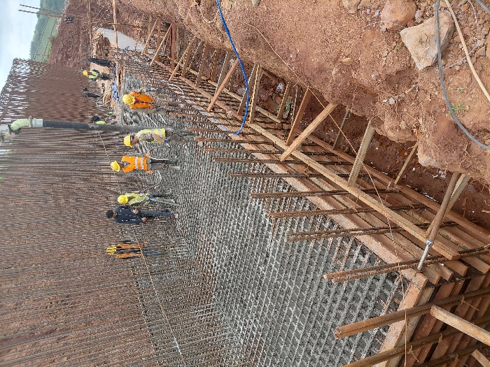 This image depicts an active construction site in Fdalate, Morocco, focused on the reinforcement and preparation for concrete pouring of a substantial foundation or structural element.
The scene is dominated by a dense grid of steel reinforcement bars (rebar) forming a rectangular base and extending vertically upwards along the perimeter, indicating the skeletal structure for a large concrete pour. Approximately ten to twelve male construction workers are visible. Six workers are centrally positioned on the rebar grid. Most workers wear yellow hard hats and high-visibility vests (orange or yellow); however, one worker in the center-left foreground wears a dark blue jacket without a visible hard hat or vest.
A large, light-green concrete pump pipe extends vertically from the upper right, with its spout positioned over the rebar grid, suggesting concrete delivery is in progress or imminent. Workers are observed standing on the rebar, some holding tools resembling rakes or vibrators, appearing to supervise or guide the concrete placement.
Along the right side of the excavated area, wooden formwork (shuttering) is installed, braced with diagonal supports against the reddish-brown soil of the excavated trench. The surrounding environment consists of disturbed reddish-brown earth from the excavation, with faint green vegetation visible on distant hills under an overcast sky. Cables and hoses are scattered on the ground near the formwork.