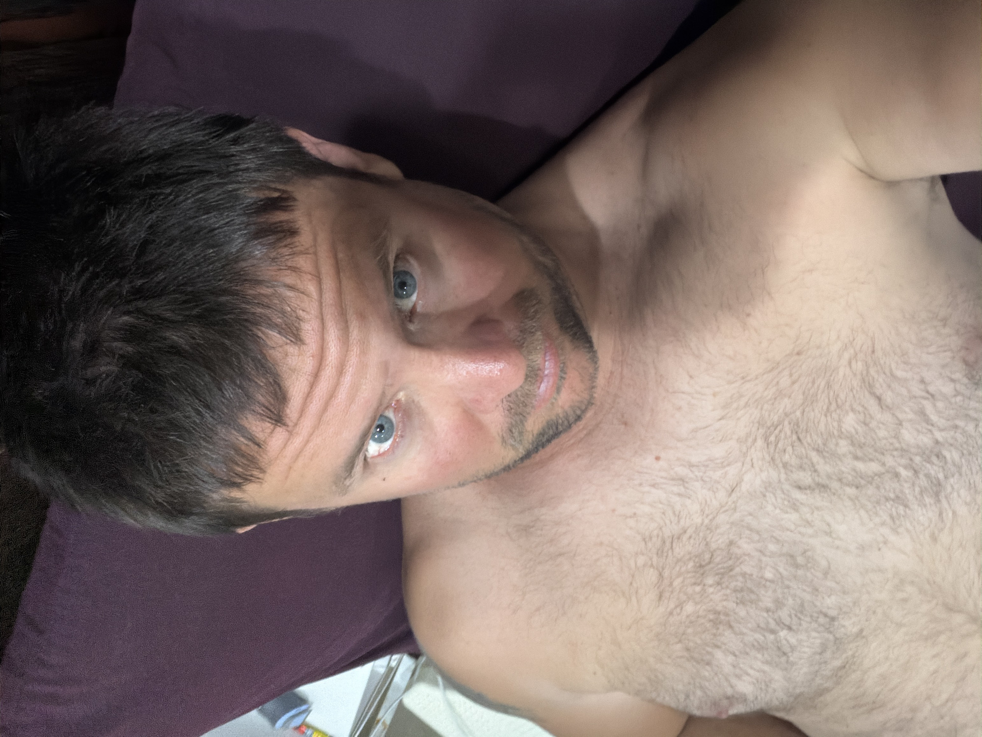 A shirtless man with short, dark hair and blue eyes is lying on his back, looking up at the camera. He has a light beard and chest hair. The background is a dark purple surface, possibly a pillow or blanket, and a portion of a white object is visible to the left. The lighting suggests an indoor setting, possibly a bedroom. There is no indication of the city of Baan Mae Nam, Thailand.