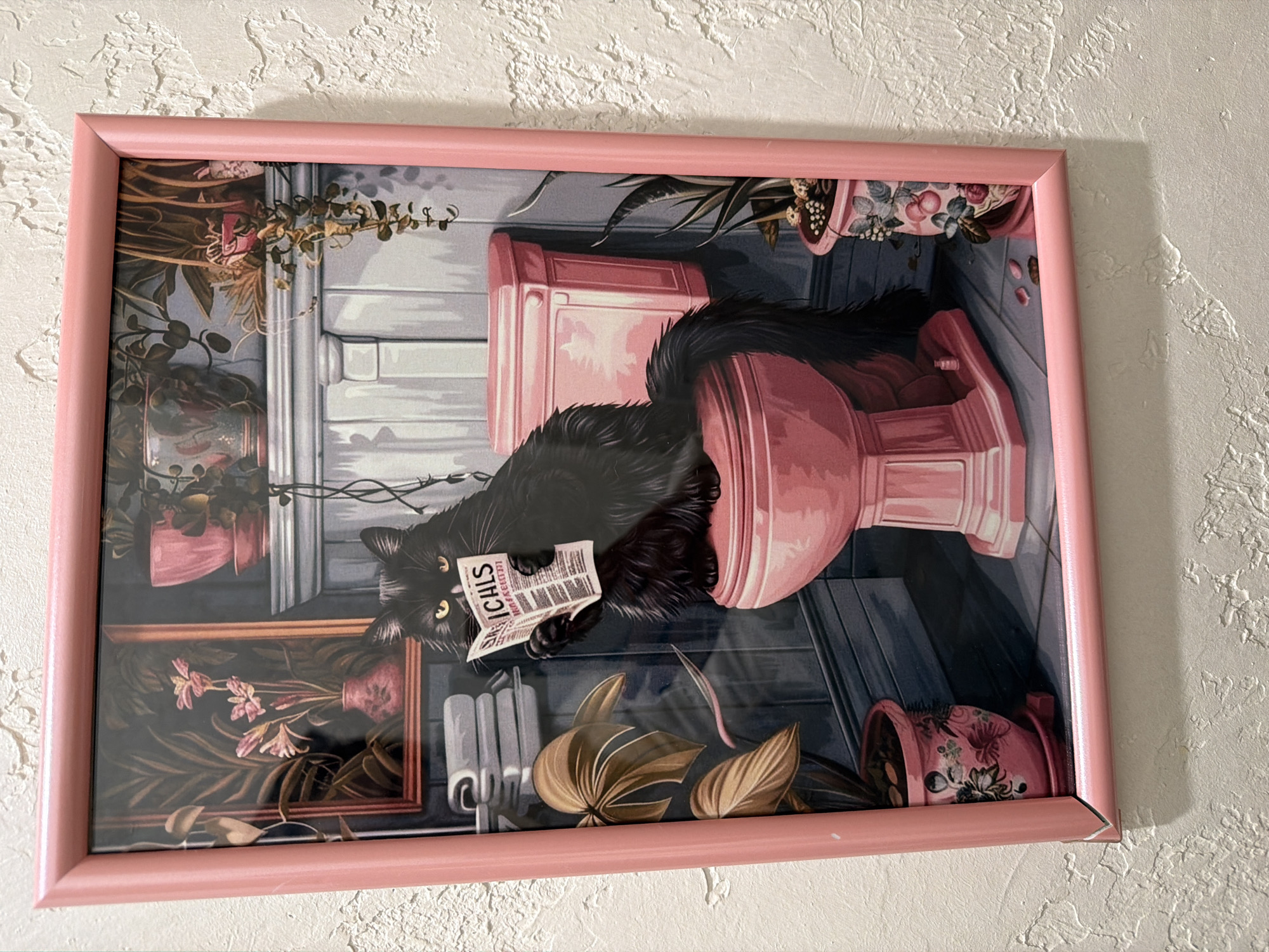 A framed print hangs on a textured, off-white wall. The print depicts a cartoon-style scene of a black cat sitting on a pink toilet, reading a newspaper. The newspaper has the word "ICHILS" printed prominently on the front page. The background includes plants, a framed picture, and other toilet-related items, all in a stylized, colorful manner. The frame itself is a light pink color. There is no evidence to suggest a specific location in Votkinsk, Russia. The image is a close-up of the framed print and does not reveal any additional contextual information about the surrounding environment.