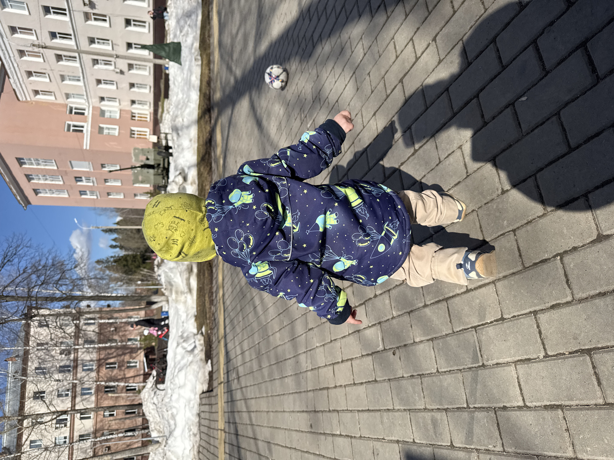 A child is playing with a soccer ball in Izhevsk, Russia. The child, wearing a yellow hat and a navy blue jacket with a pattern of space vehicles, stands on a paved walkway. The walkway is made of rectangular concrete pavers and is partially shaded. The soccer ball is slightly ahead of the child. In the background, there are residential buildings and patches of snow. A military tank is visible behind one of the buildings. Bare trees line the area behind the walkway.
