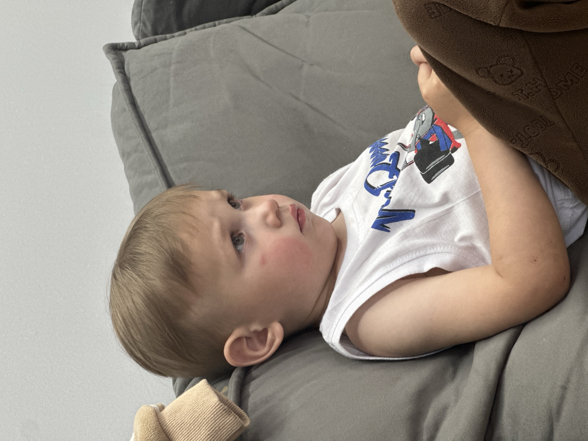 A young child is lying on a gray sofa. The child is wearing a white t-shirt with a cartoon graphic and has blonde hair. Part of a brown blanket or garment is visible on the right side of the frame. The background includes a light gray wall. This image was taken in an indoor setting. The location in the city of Эстачи, Russia, is not apparent from the image.