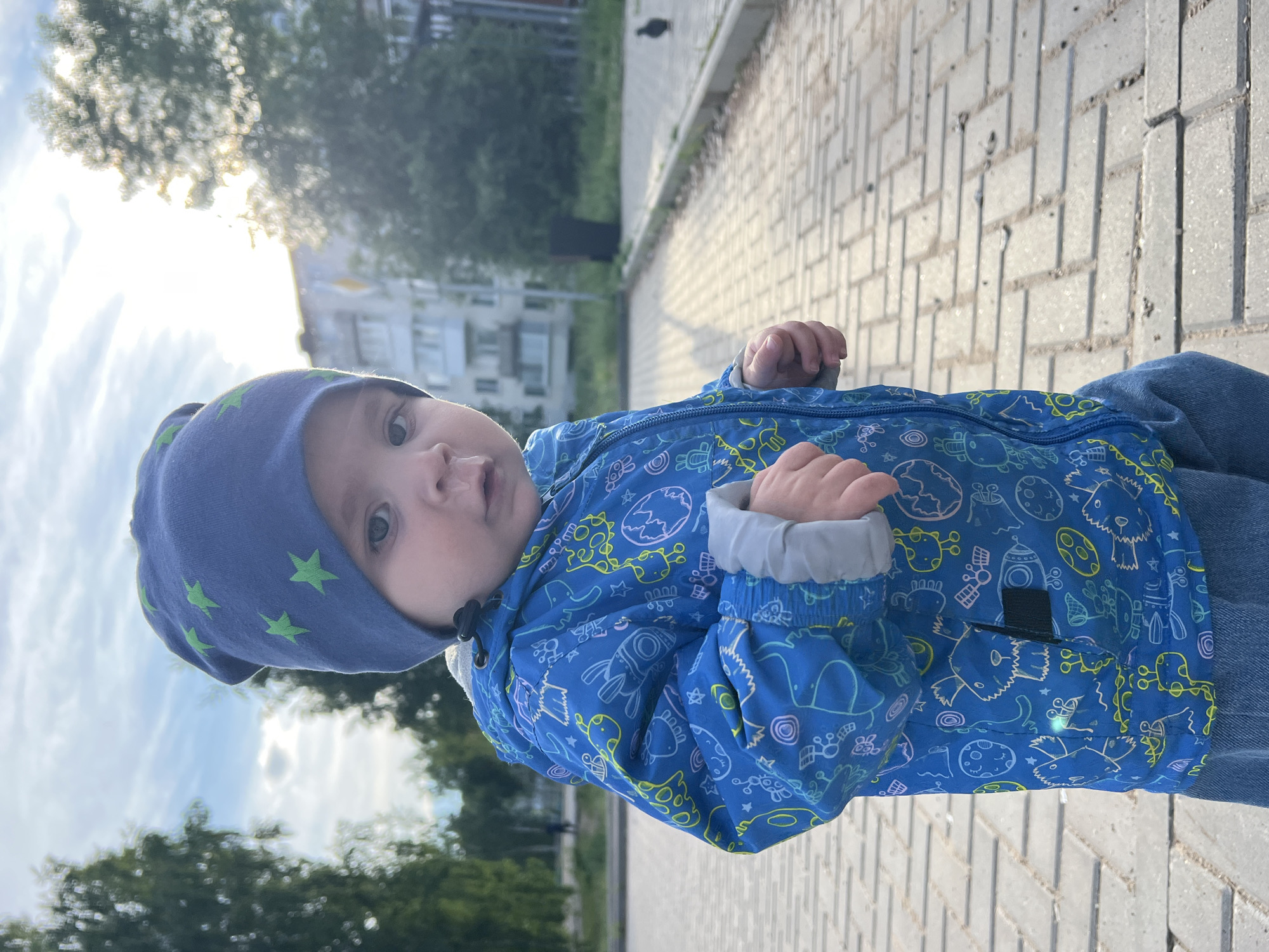 A toddler is standing on a brick walkway in a public area. The child is wearing a blue jacket with cartoon drawings, a blue hat with green stars, and jeans. Buildings and trees are visible in the background. The location is Noviy, Russia.