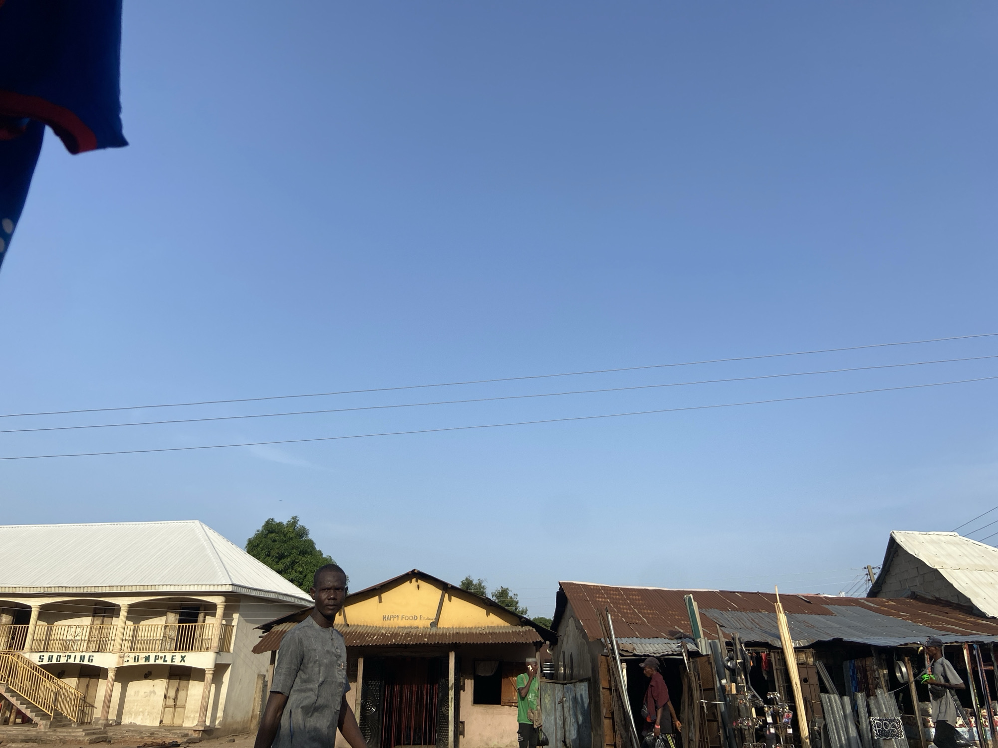The image depicts a street scene in Mararaba, Nigeria. A man wearing a grey shirt walks past a two-story building labeled "SHOPPING COMPLEX" with a white roof and yellow stair railings. Next to it is a one-story yellow building with the words "HAPPY FOOD" on its facade. Adjacent to that, a rusty corrugated metal structure contains various items, with two people present. One wears a green shirt, and the other is in a red shirt. Power lines are visible against a clear blue sky.