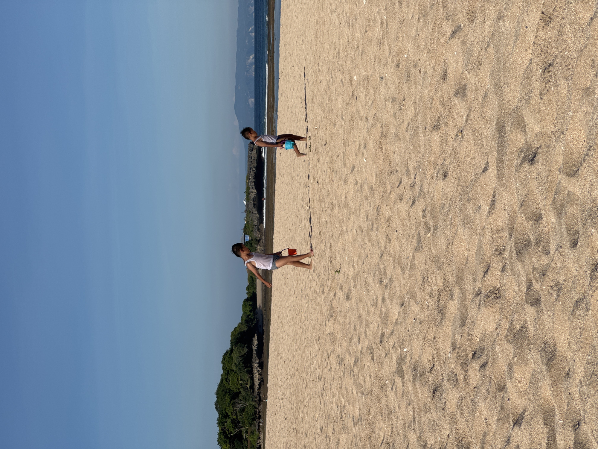 Two children are on a sandy beach in Kuta Selatan, Indonesia. The sky is blue with a slight haze near the horizon. The shoreline is visible in the background, with a narrow strip of vegetation separating the beach from the ocean. The sand is light brown and textured. One child is holding a red bucket while the other child is holding a blue bucket. Both are facing towards the ocean.