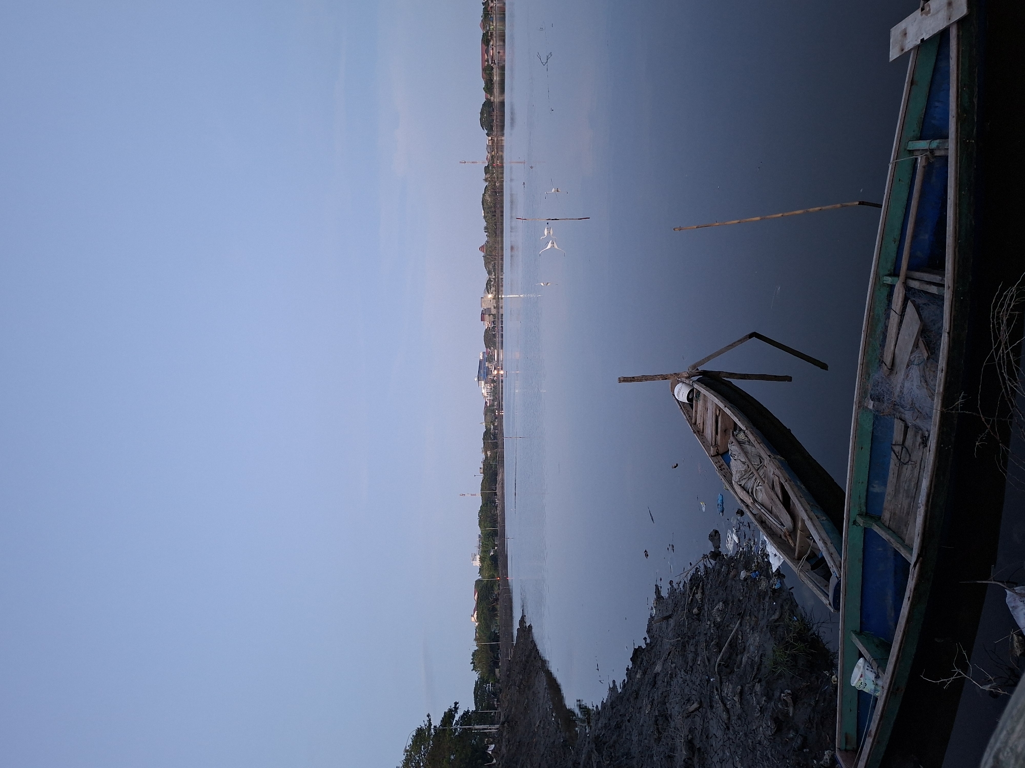 The image shows a view of a body of water at dusk. In the foreground, there are two boats, one with green and blue trim. The shoreline is muddy. Across the water, there is a visible skyline of a city with lights, possibly Lhokseumawe, Indonesia. There are several birds flying above the lake.