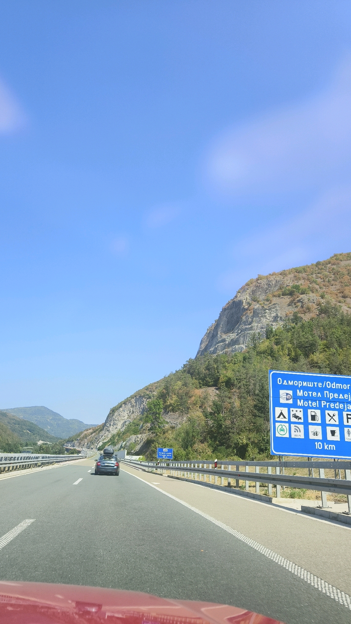The image captures a highway scene in the Vladicin Han Municipality, Serbia, under a clear blue sky. The highway stretches into the distance, flanked by metal guardrails. On the right side of the road, a blue directional sign indicates "ОДМОРИШТЕ/ODMORIŠTE" and "Motel Predejane" with an arrow pointing ahead, along with icons for camping, hotel, gas station, restaurant, and parking, specifying "10 km". In the foreground, the hood of a red car is visible. A gray car with a roof rack is driving ahead on the highway, and further in the distance, other vehicles can be seen. The landscape features a steep, rocky mountain slope on the right, covered in sparse vegetation and trees. On the left side, rolling hills with trees and greenery recede into the background. A smaller blue sign is visible further down the highway on the left. The overall impression is of a scenic highway route through a mountainous region.