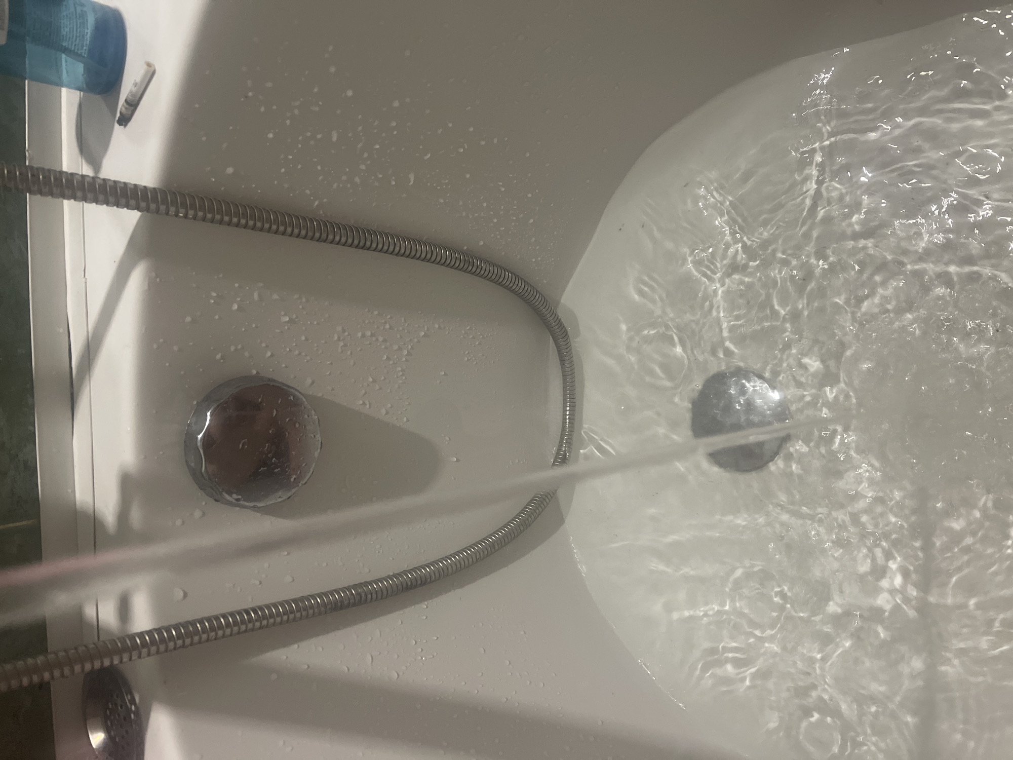An overhead shot captures a white bathtub actively filling with water. A silver, flexible shower hose is coiled within the tub, with a strong stream of water flowing from an unseen shower head (presumably connected to the hose) into the basin, creating significant ripples and movement on the water's surface. A chrome drain cover is submerged in the water, reflecting the light.
On the left side of the tub, a second chrome fixture, likely an overflow or another drain, is visible, covered in water droplets. Resting on the tub's edge in the upper left corner is a blue plastic bottle, possibly containing shampoo or shower gel, with faint, unreadable text on its label. Adjacent to the bottle, a small, light-colored cylindrical object, possibly a cigarette butt or similar debris, is present. The white surface of the bathtub is speckled with water droplets, indicating a wet environment. The scene is indoors, illuminated by artificial light, and no people are visible. This setting is located in Poltava, Ukraine.