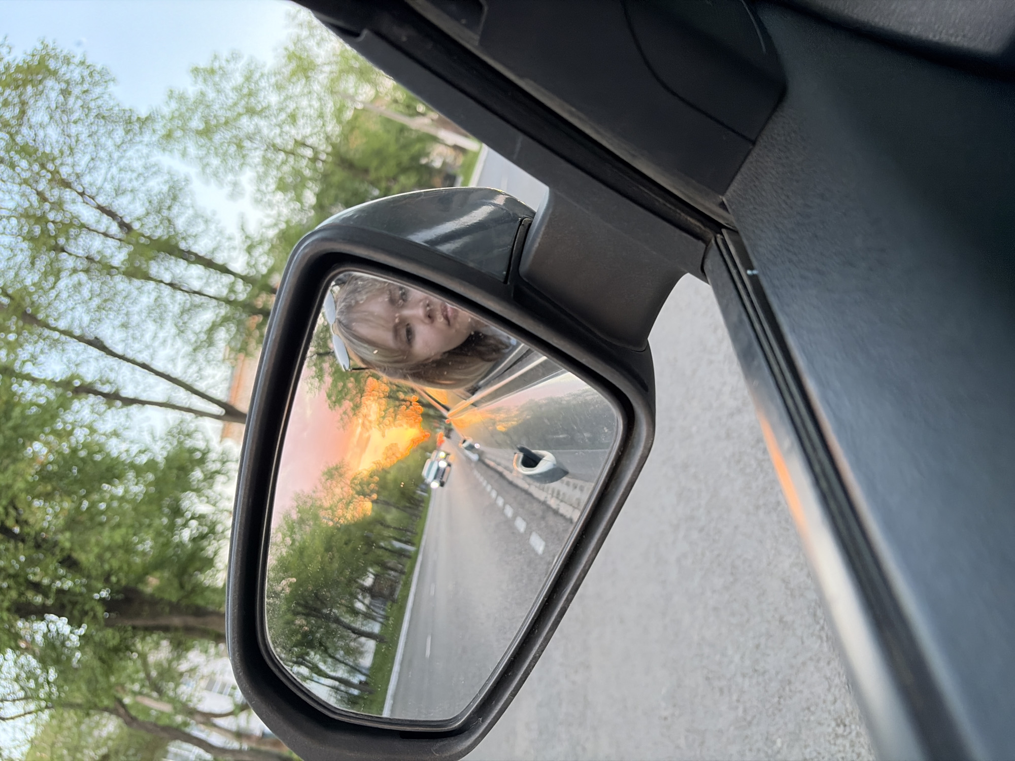 The image shows the side mirror of a vehicle. The reflection in the mirror reveals a woman with blonde hair looking into the mirror. Behind her, the reflection shows a road lined with trees, a car further down the road, and a vivid sunset visible in the background. Outside the mirror, trees are visible, as well as the interior of the car. The location is likely Chaikovsky, Russia, as specified in the prompt.