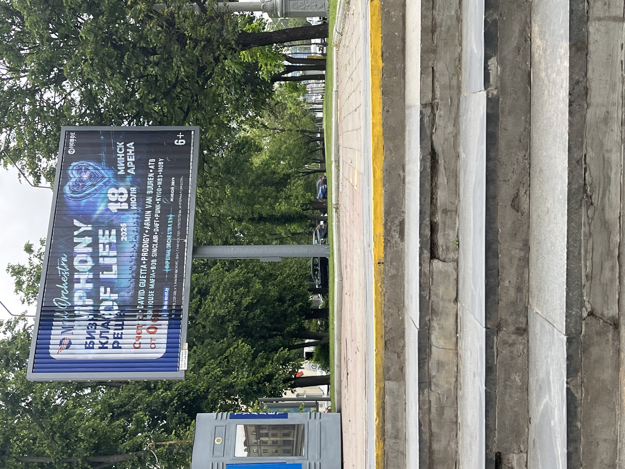 The image displays a daytime scene in Minsk, Belarus, featuring a large, partially rotating slat billboard prominently positioned above a street. In the immediate foreground, a set of four weathered concrete or stone steps with dark grey treads and lighter grey risers ascends to a red-paved pedestrian area edged with a yellow curb.
The billboard, supported by a grey pole, is divided into two distinct advertisements. The left portion, partially obscured and distorted by the rotating slats, features a blue and white design with text in Cyrillic, including "БИЗНЕС КЛАСС" (Business Class), "Счет от 0" (Account from 0), and "ООО «Бизнес»" with a phone number "+375(29)11-44-666". The right portion, set against a dark, cosmic-themed background with blue and purple hues, advertises "IMPERIAL ORCHESTRA: SYMPHONY OF LIFE" for July 18, 2026, at "МИНСК АРЕНА" (Minsk Arena), marked with a "6+" age restriction. A stylized heart graphic with circuit patterns is visible, along with a list of performing artists including David Guetta, Prodigy, Armin van Buuren, ATB, Swedish House Mafia, Bob Sinclair, Daft Punk, Kygo, Moby, and M33. The text "ЖИВОЙ ЗВУК" (Live Sound) and a phone number "+375(29)220328" are also present.
Behind the red-paved area and extending into the midground, a light-colored paved walkway leads to an asphalt road. Both sides of the road are lined with a dense arrangement of mature deciduous trees with lush green foliage, forming a boulevard. Several dark-colored cars are parked along the road, and a light-colored vehicle is partially visible between trees further down the road on the left. On the far left, a grey utility box or advertising stand reflects a distant building. The sky above is overcast. No individuals are visible in the scene.