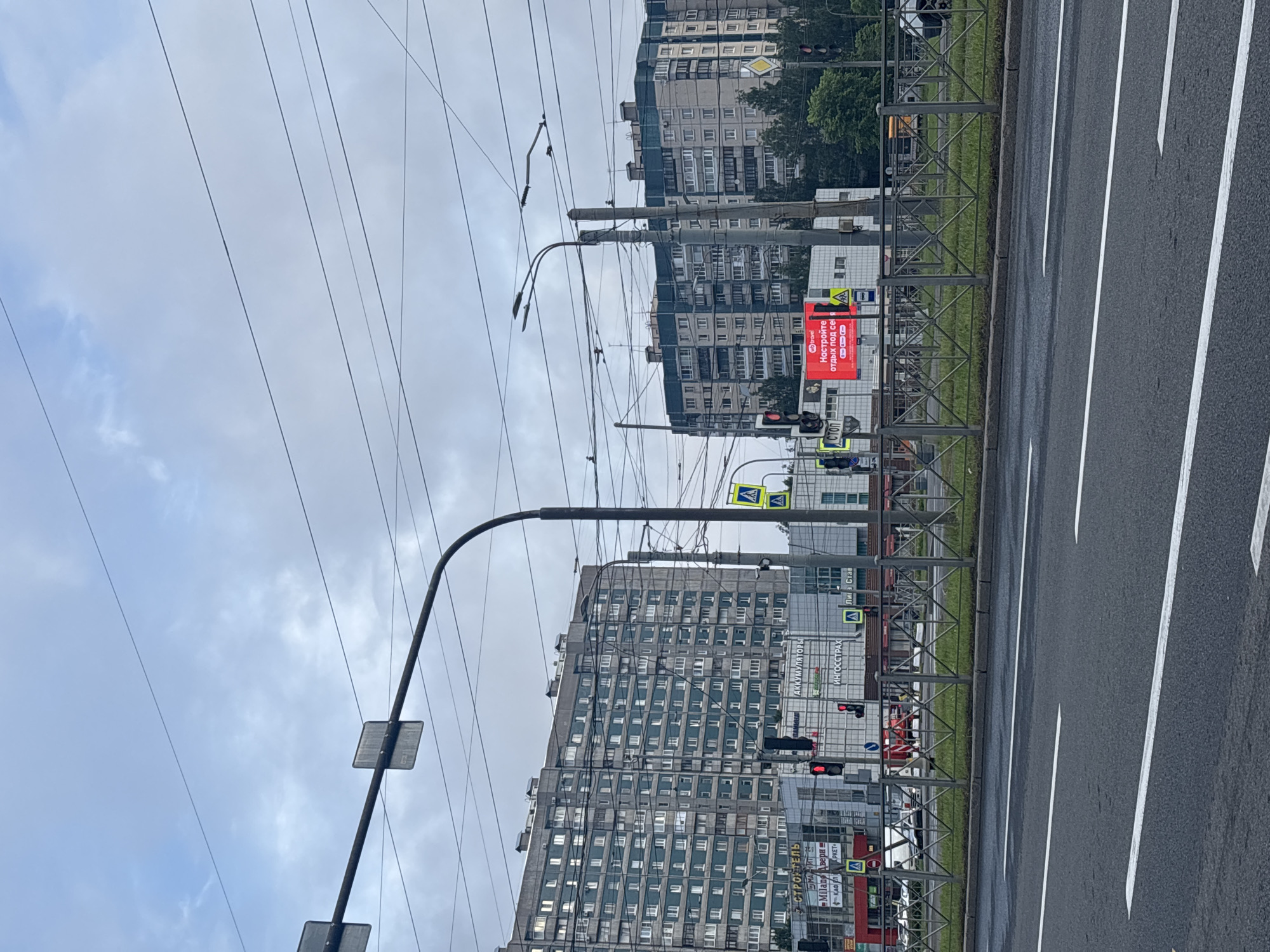The image shows a street scene in Saint Petersburg, Russia, under a cloudy sky. In the foreground, there is an asphalt road with white lane markings and a metal guardrail separating it from a grassy area. Behind the guardrail, several buildings are visible, including tall, multi-story apartment buildings with many windows. Some of these buildings appear to be older, Soviet-era architecture. In the midground, there are commercial buildings with signage, including one that reads "АККУМУЛЯТОРЫ" (Batteries) and another that displays a red advertisement with Russian text and a logo that says "wttravel". Traffic lights are visible, some showing red. Overhead, a complex network of electrical wires crisscrosses the sky, supported by poles. A traffic sign with an exclamation mark inside a yellow triangle is also present. The overall impression is of a typical urban environment in Saint Petersburg on an overcast day.