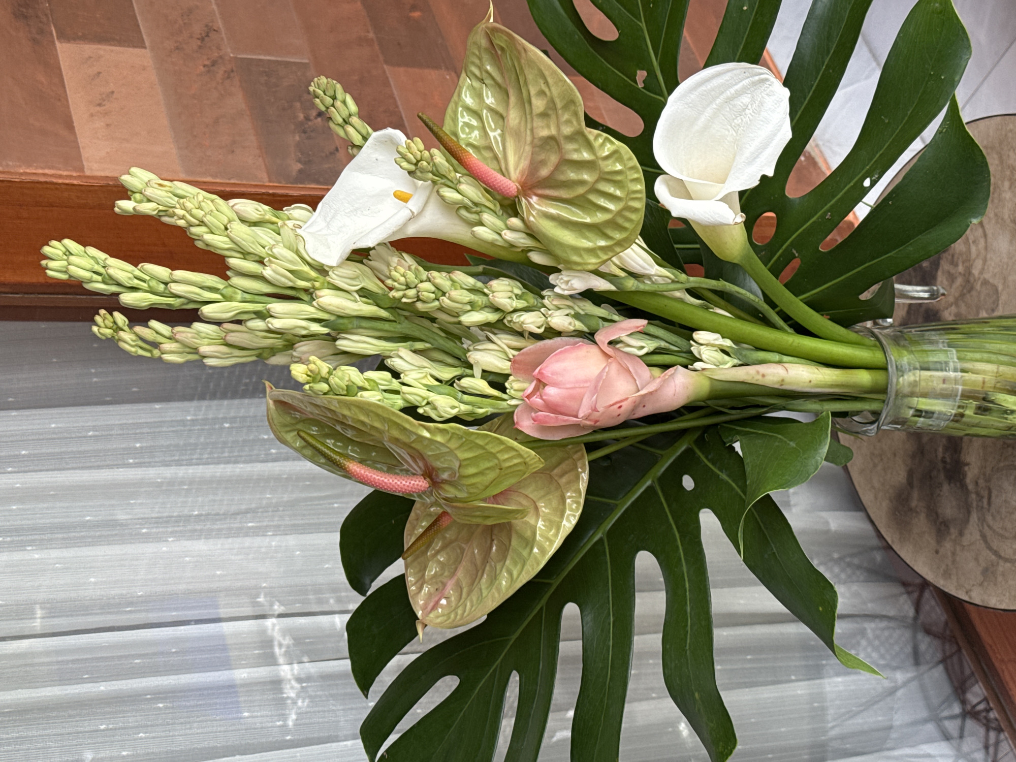 The image shows a bouquet of flowers, including white calla lilies, green anthuriums, tuberoses, and a pink ginger flower, arranged with a monstera leaf. The bouquet is likely in a glass vase. The background includes a wooden surface, a set of white blinds, and a glimpse of an outdoor environment. The location is identified as Mengwi, Indonesia, based on provided information. No people are visible in the image.
