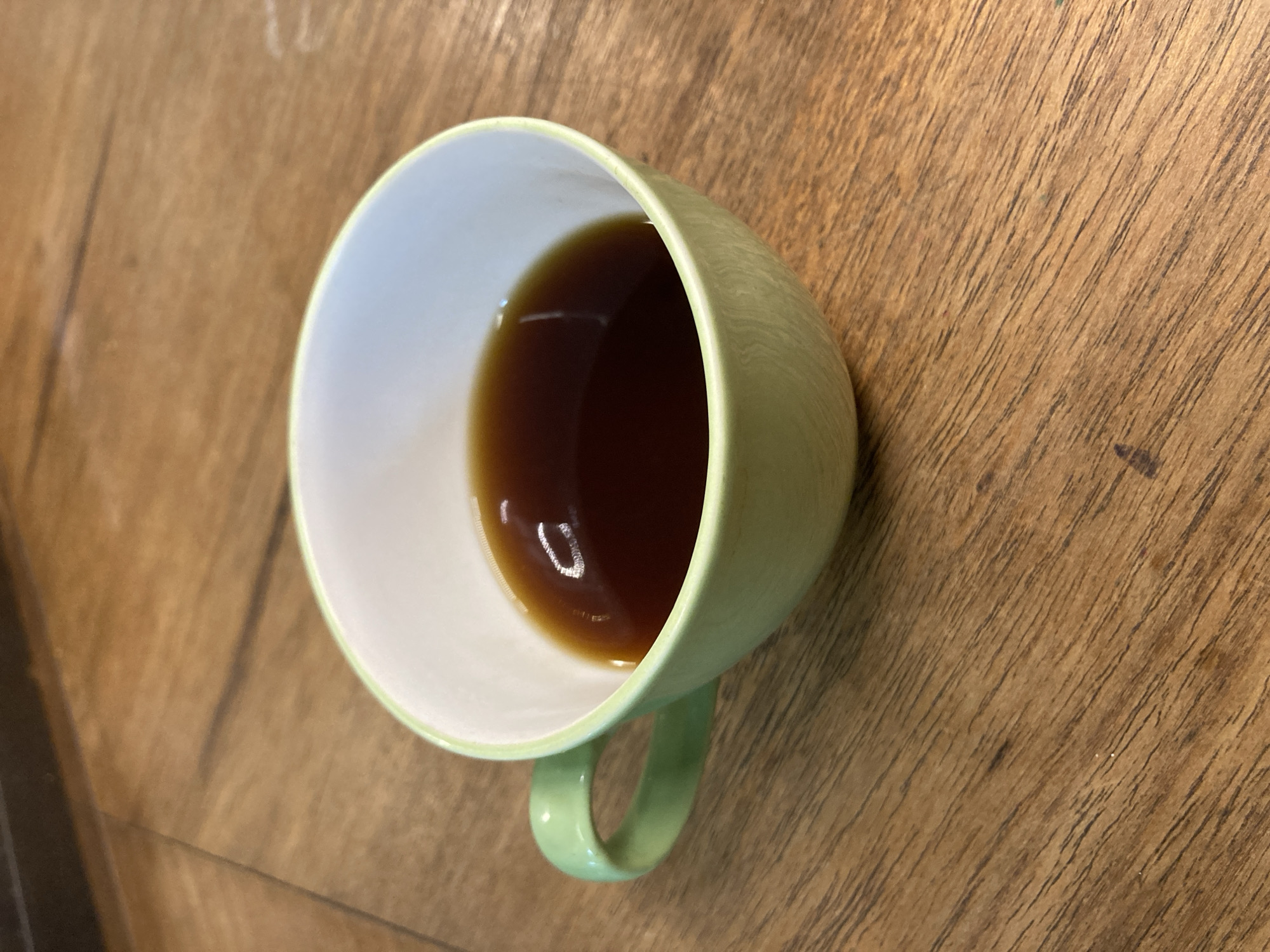 The image displays a light green mug with a white interior, containing a dark brown liquid, likely coffee or tea. The mug is positioned on a wooden surface, which exhibits clear grain patterns and variations in tone. A bright, oval-shaped reflection is visible on the surface of the liquid within the mug, indicating an overhead light source. The handle of the mug is visible on the lower side. The photograph is taken from a top-down perspective, focusing on the mug and its immediate surroundings. The setting appears to be indoors, possibly on a table or counter. There are no people, discernible activity beyond the presence of the mug, or visible text in the image.