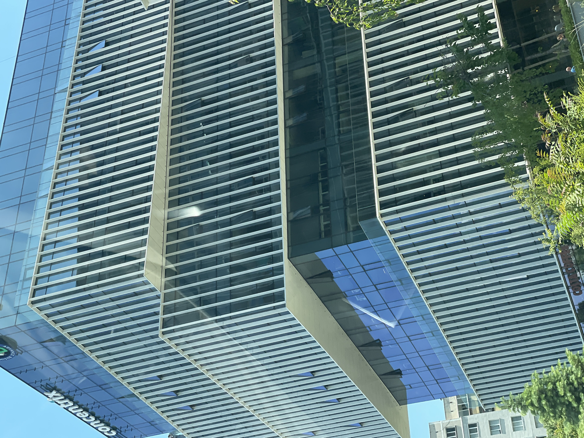 This is a medium-angle, exterior shot of the Concentrix building in Sofia, Bulgaria. The modern glass and steel structure features a staggered design with prominent vertical white fins. Sunlight reflects off the glass facade. In the foreground, lush green trees partially obscure the lower levels of the building. A residential building can be seen to the left. A few people are visible near the bottom of the frame, on a sidewalk. The sky is clear and blue.