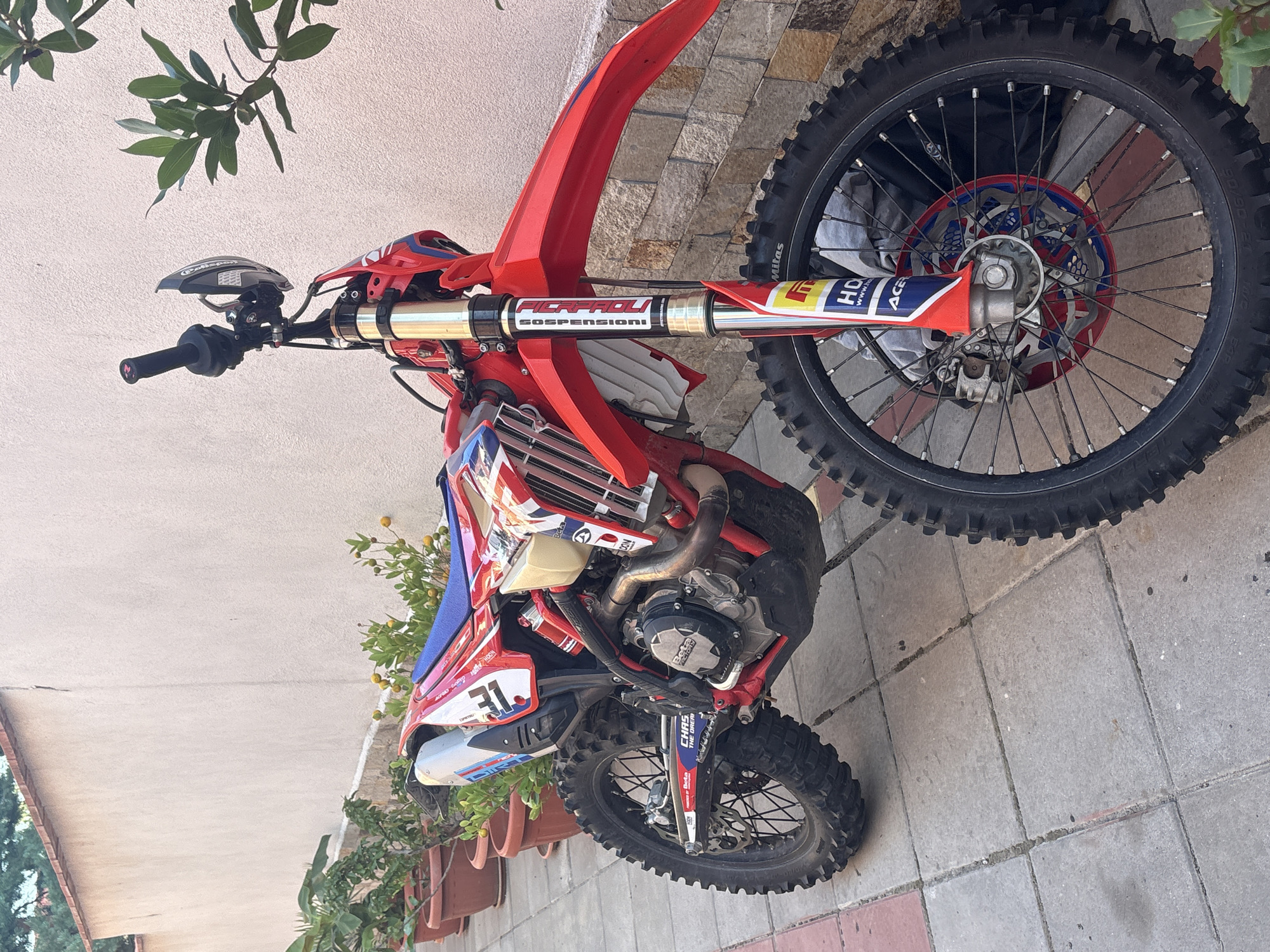 A red and white dirt bike is parked on a paved patio next to a cream-colored wall in Strelcha, Bulgaria. The motorcycle has black tires with knobby treads, silver spokes, and a red front fender. It features numerous sponsor decals, including "Beta" and "Mitas". Several potted plants with green leaves and some small yellow fruits are arranged along the base of the wall. The scene appears to be outdoors in daylight.