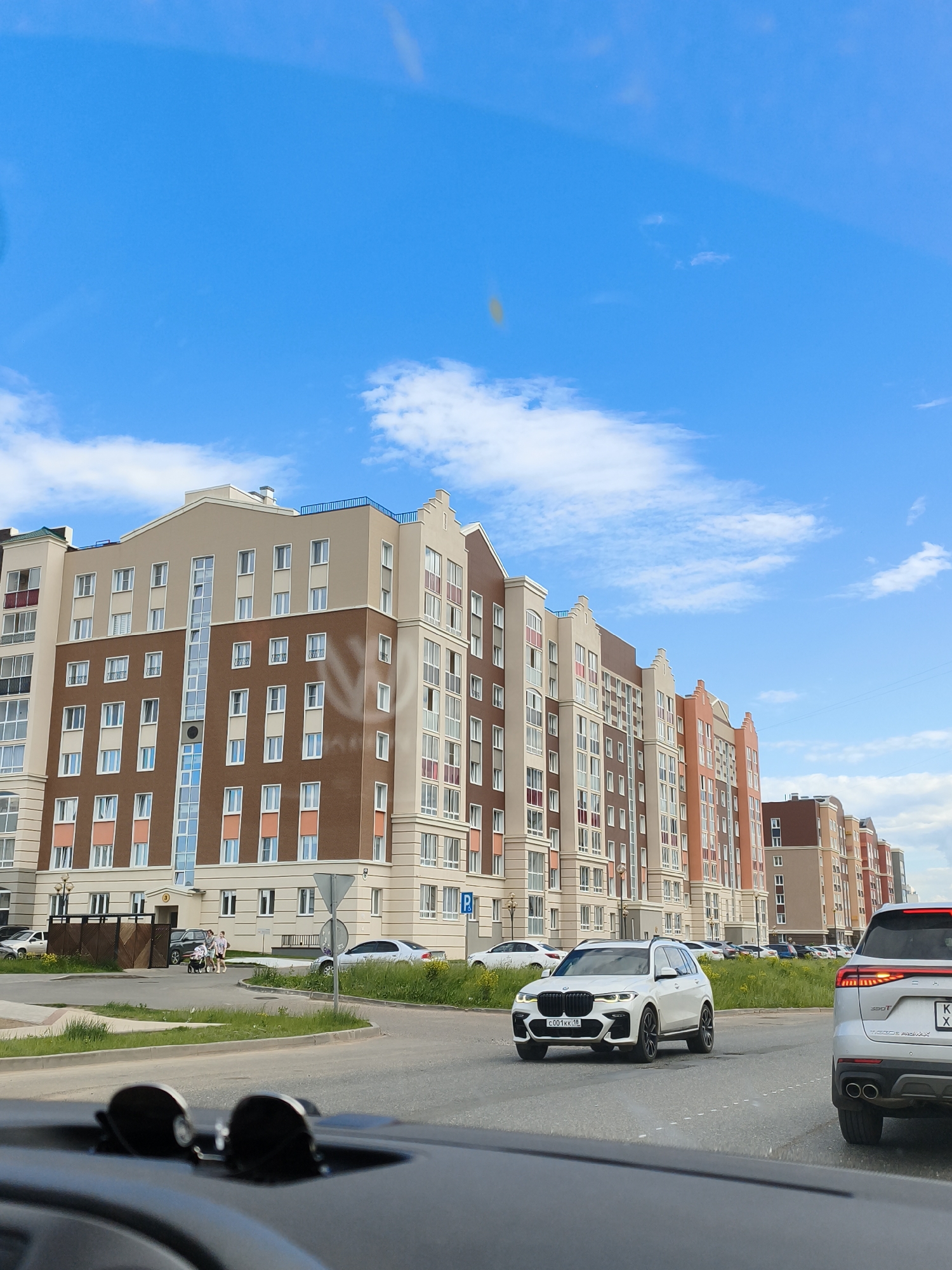 In Izhevsk, Russia, a multi-lane paved road runs parallel to a row of modern, multi-story apartment buildings. The buildings feature a mix of brown and beige facades with numerous windows and balconies. The sky is bright blue with scattered white clouds. On the road, several cars are visible. In the foreground, a white BMW SUV is driving towards the viewer, and further ahead, a silver car is also moving. In the background, pedestrians are walking near the apartment buildings. The scene appears to be a residential area within the city.