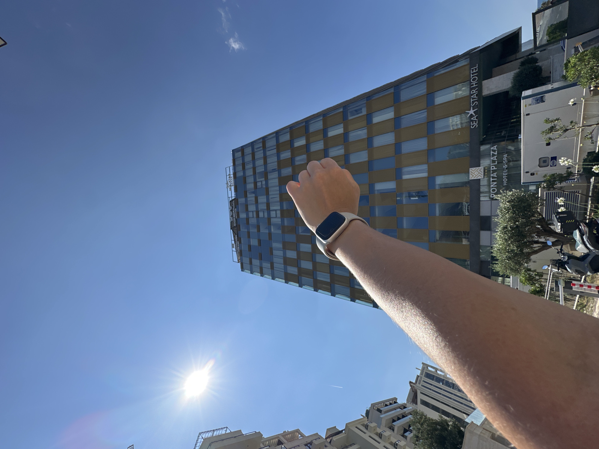 An arm wearing a smartwatch is extended towards a tall, modern hotel building in Boreti, Montenegro. The hotel, named "SEA STAR HOTEL" and "PONTA PLAZA HOTEL & SPA," has a facade of gold and blue glass panels. The sky is a clear, bright blue, with the sun shining brightly and creating a lens flare effect. In the background, other buildings and trees are visible. The arm appears to be positioned to frame or point at the hotel.