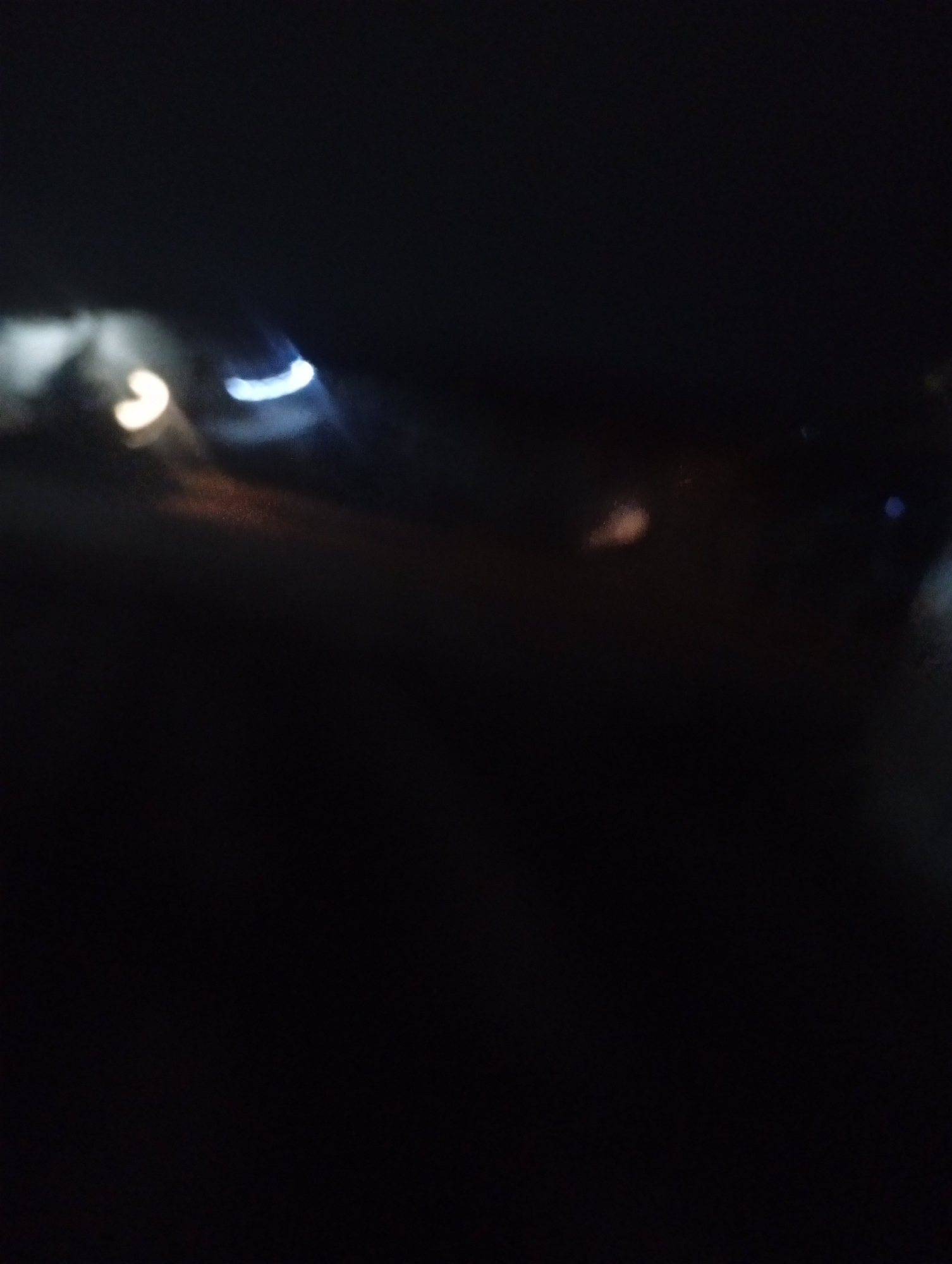 The image depicts an out-of-focus scene, likely at night, with several blurred lights. In the upper left quadrant, a bright, yellowish light with a curved shape is visible. To its right, a bright, bluish-white curved light is present. Further to the right and slightly lower, a fainter, reddish-brown blurred area suggests another light source. The rest of the image is predominantly black, indicating a dark environment. No specific landmarks or recognizable objects are clearly discernible due to the lack of focus. The scene does not provide enough detail to determine its location within Bukuru, Nigeria, or to identify any people, subjects, actions, or interactions.