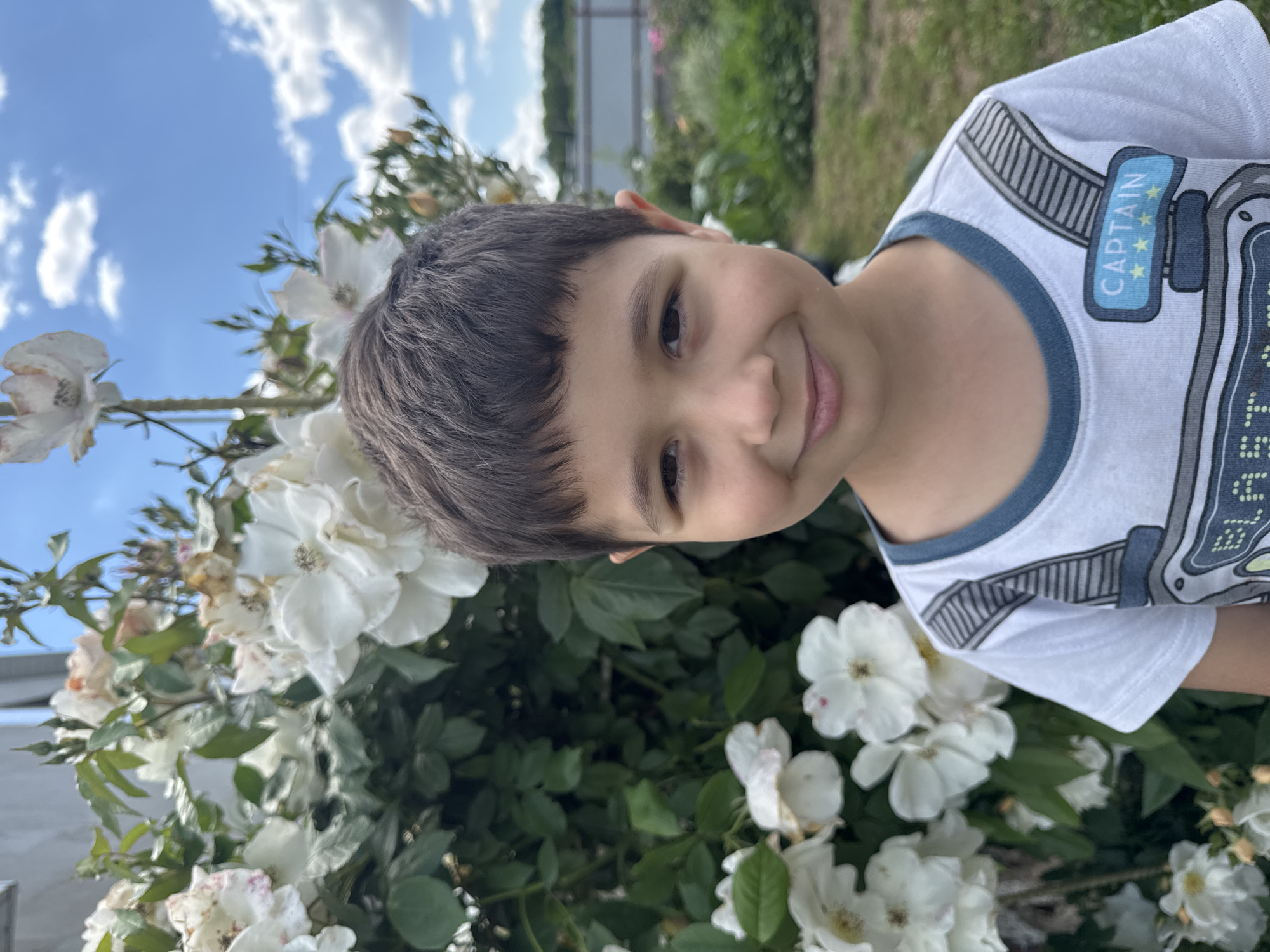 A young boy smiles at the camera, standing in front of a bush with white roses. He is wearing a white t-shirt with a blue collar and a graphic design that includes the word "CAPTAIN" and three stars. The background shows a fence and green foliage, with a blue sky and white clouds above. The image appears to have been taken outdoors in daylight.