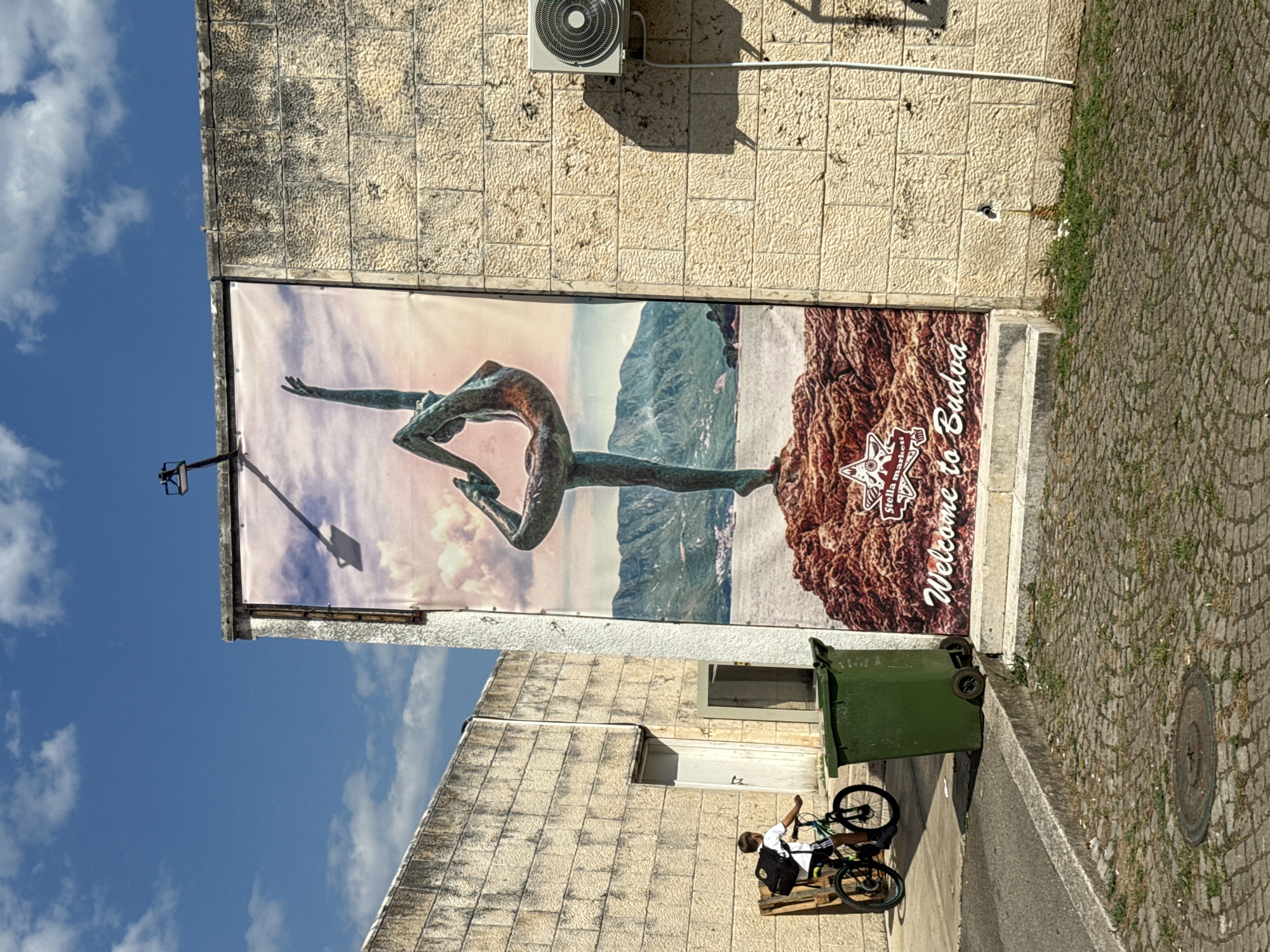 The image depicts a street scene in Budva, Montenegro, featuring a large banner on a stone building. The banner displays a statue of a dancer in a yoga pose against a backdrop of mountains and water, with the text "Welcome to Budva" at the bottom. To the left of the banner, a young boy on a bicycle wearing a backpack is visible. A green trash bin stands next to him. The buildings are made of light-colored stone blocks, and the ground is paved with cobblestones. The sky is blue with scattered clouds. An air conditioning unit is mounted on the wall to the right of the banner.