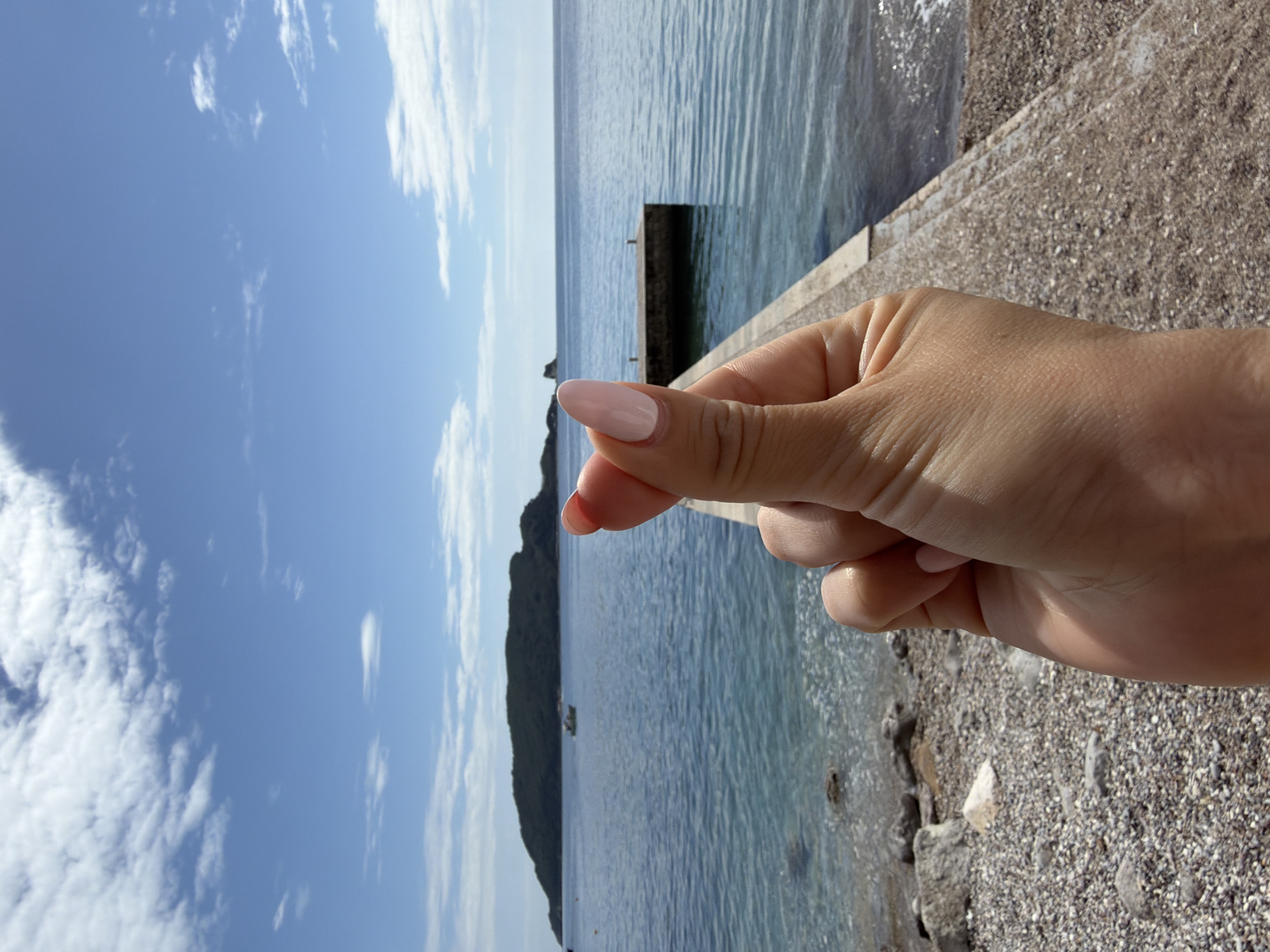 The image shows a hand making a heart shape against a seascape backdrop. The hand, with light-colored nail polish, is positioned in the foreground. Behind it, the Adriatic Sea stretches out, with a small, rocky island visible in the distance. The sky is blue with scattered clouds. A pebbly beach and a stone walkway are visible in the foreground, suggesting a coastal setting. Based on the general scenery of the Adriatic Sea with rocky outcrops and islands, and the urban coastal elements, this is possibly a shot taken at or near Budva, Montenegro. A structure extends into the water near the hand.