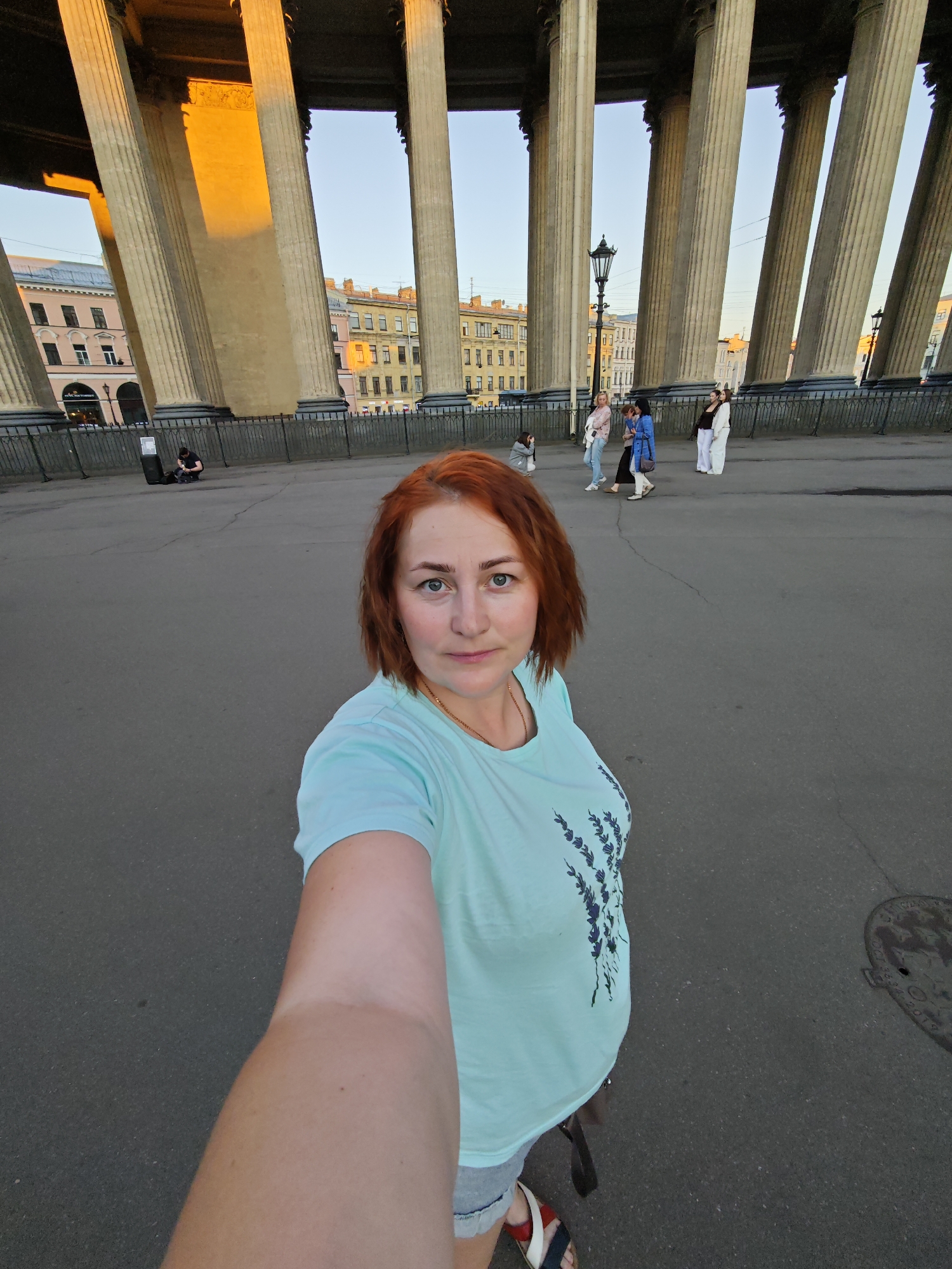 The image captures a selfie of a woman positioned in the foreground, standing on a paved surface in front of the Kazan Cathedral colonnade in Saint Petersburg, Russia.
The woman in the foreground has reddish-brown, medium-length hair and light-colored eyes. She is wearing a light mint-green short-sleeved t-shirt adorned with a dark blue, stylized floral or branch pattern on the left side of the chest, light-colored denim shorts, and sandals with red, white, and blue straps. Her right arm, extended forward, is prominent in the lower right portion of the frame, holding the camera. She maintains a neutral facial expression, looking directly at the camera. A thin gold chain is visible around her neck.
The mid-ground features a dark gray paved area with noticeable cracks. To the left, a person dressed in dark clothing is crouched next to a dark, rectangular object resembling a speaker or amplifier. Further right, a group of five individuals is visible. One person wears a light blue long-sleeved shirt and light-colored pants. Another has dark hair and wears a blue jacket over dark clothing. A third individual, with dark hair, wears a white shirt and dark pants. Two women stand together on the right: one in an all-white outfit and the other in a dark top with white trousers. A black, ornate street lamp with a single lantern stands upright in the mid-ground.
The background is dominated by the colossal, fluted stone columns of the Kazan Cathedral's colonnade. The columns exhibit a beige and light gray coloration. Direct sunlight, appearing golden, illuminates the upper portions and inner surfaces of some columns, indicating a low sun angle. Beyond the colonnade, several multi-story buildings with facades in shades of pink, yellow, and brown are visible. A clear, light blue sky is visible above and between the columns. A dark, wrought-iron fence runs along the base of the colonnade, separating it from the paved area. A circular metal manhole cover is partially visible on the far right foreground, near the woman's foot.
