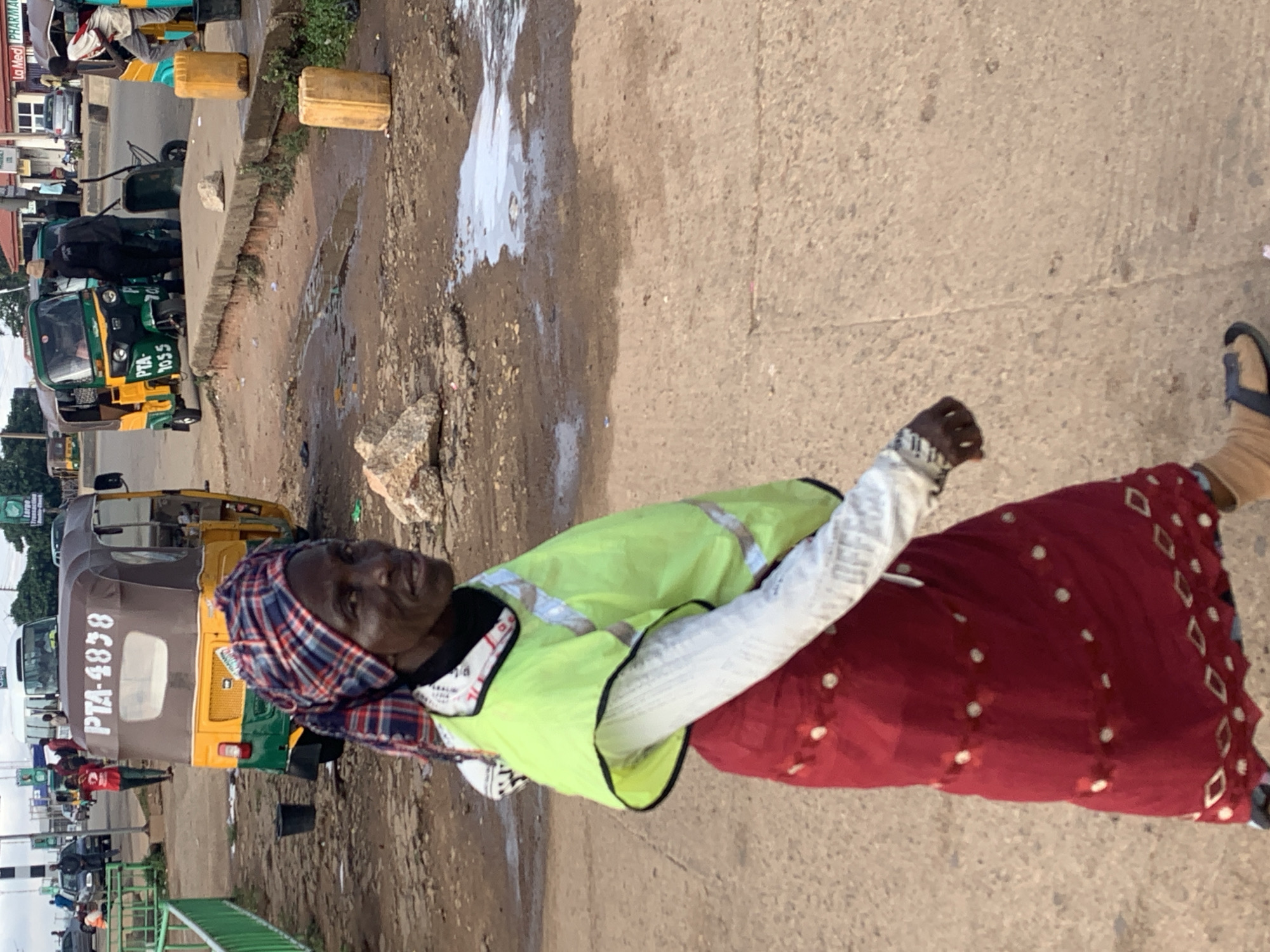 A woman walks in the foreground, facing right, on a concrete pathway. She wears a red skirt with white circular patterns and a contrasting design at the hem, a white long-sleeved shirt, and a bright lime green vest with reflective stripes. Her head is covered with a red, blue, and white checkered headwrap. Behind her, on a dusty, unpaved surface, are several three-wheeled vehicles, commonly known as Keke-Napeps, and a white bus in the background. Yellow plastic containers are visible on the ground, and some areas are wet or muddy, indicating recent rain or spills. The scene appears to be in a public area, possibly a transport hub or market, in Gura Top, Nigeria.