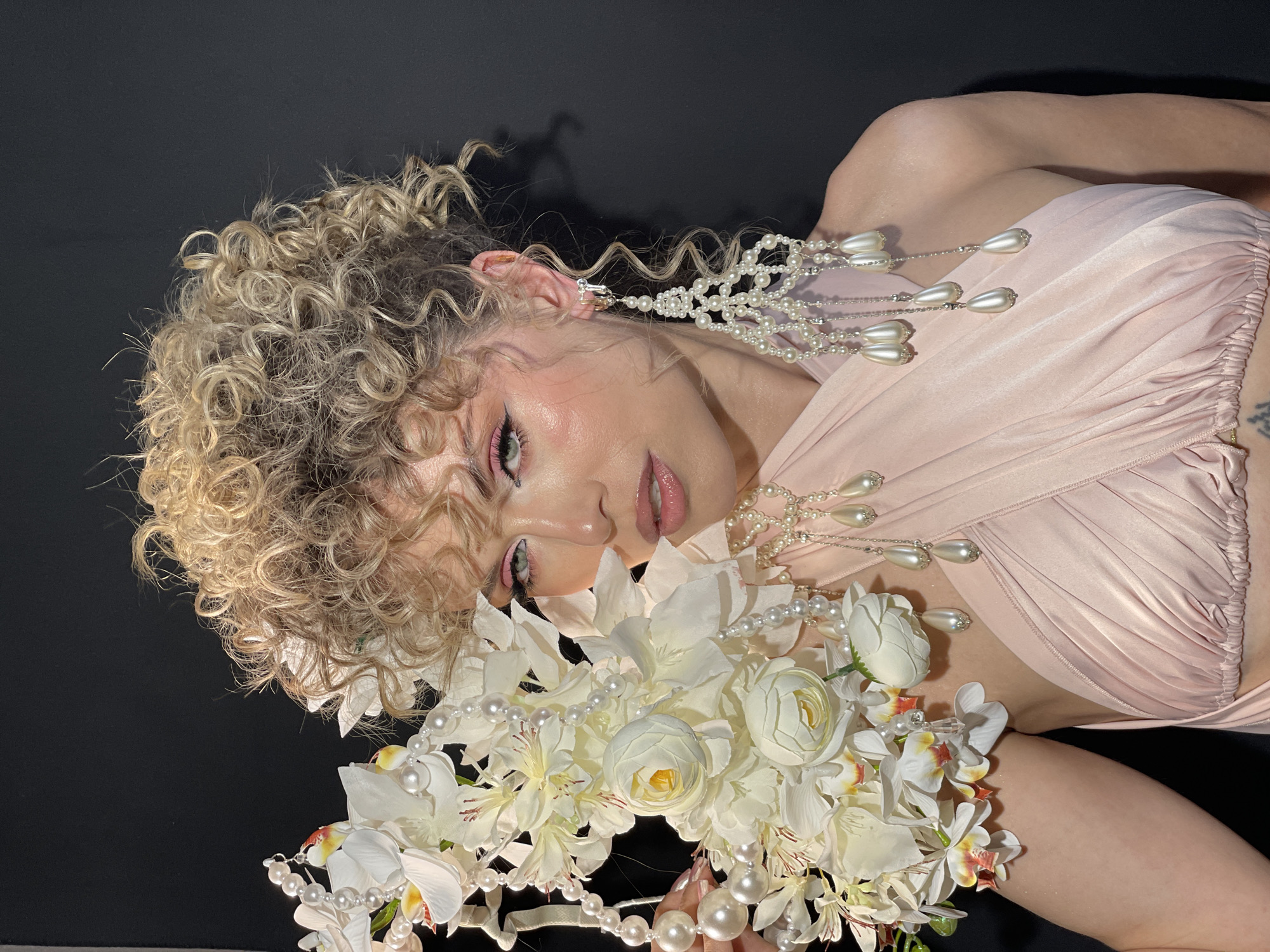 A woman with blonde curly hair is reclining against a black background. She is wearing a light pink dress. She has ornate pearl earrings and a flower crown. She is holding a bouquet of white flowers. There is no indication of the city of Kazan, Russia in the image.