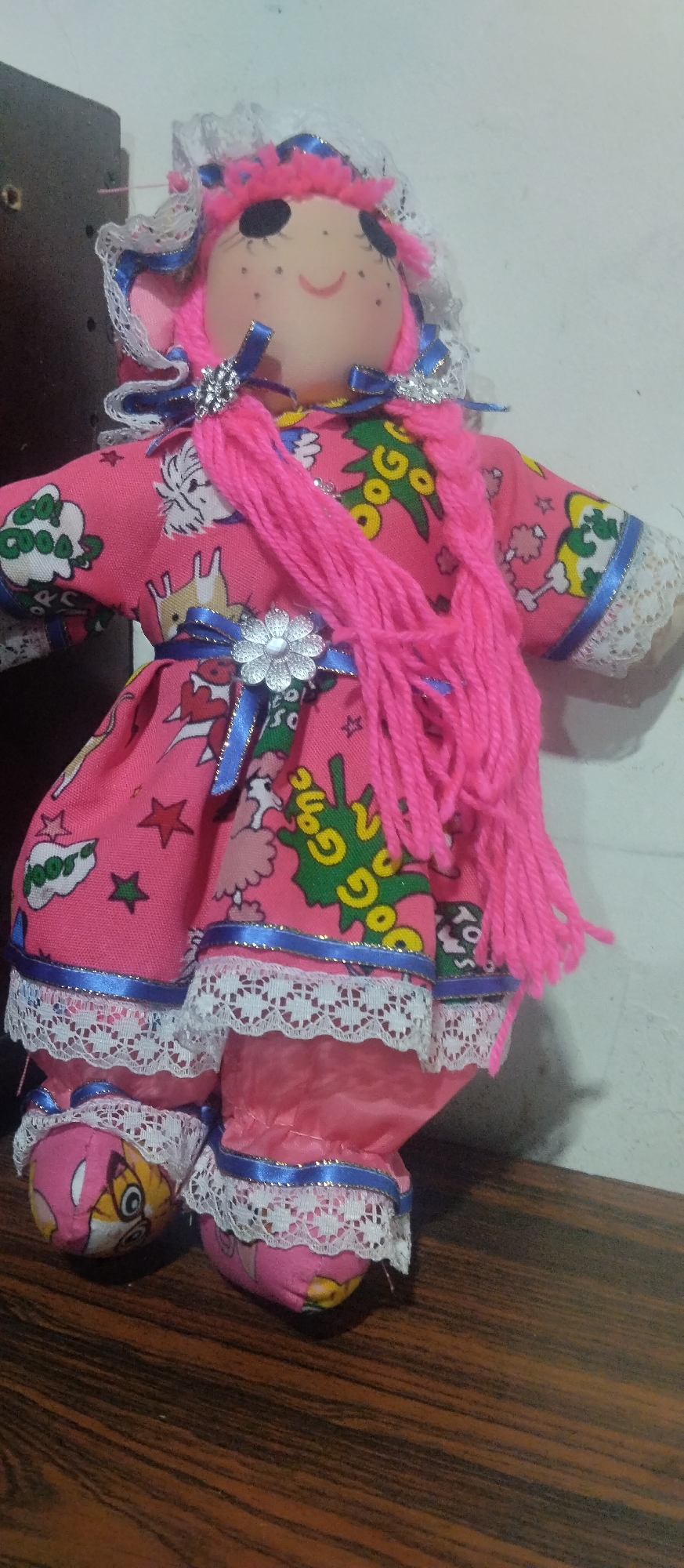 The media file showcases a brightly colored, handmade fabric doll, likely positioned on a dark wooden surface against a plain white wall in an indoor setting. The doll has a light pink face with stitched black eyes, eyelashes, light pink cheeks, and a red smiling mouth. Her hair is made of vibrant pink yarn, styled into pigtails tied with blue ribbons and silver-colored decorative pieces. She wears a white lace bonnet, also trimmed with blue ribbon.
The doll's dress is a vivid pink fabric adorned with various cartoon-like patterns. Visible text includes "Goo Goo" in green lettering, white cloud shapes with "Goos" or "Goop," and small stars. Abstract animal figures are also part of the pattern, such as a white dog-like character. The short sleeves of the dress are edged with blue ribbon and white lace. A silver-colored flower buckle secures a blue ribbon belt around her waist. The bottom hem of her dress and her fabric pants or bloomers are similarly decorated with blue ribbon and white lace. Her soft fabric "shoes" feature cartoon faces, with visible details suggesting characters like Amy Rose and Tails from the Sonic the Hedgehog series.
The doll is posed in an upright, cheerful manner, smiling. No specific activity or event is taking place; the doll appears to be on display. The lighting is even, suggesting an indoor environment. Given the location context of Caracas, Venezuela, this doll may represent a local craft or handmade item.