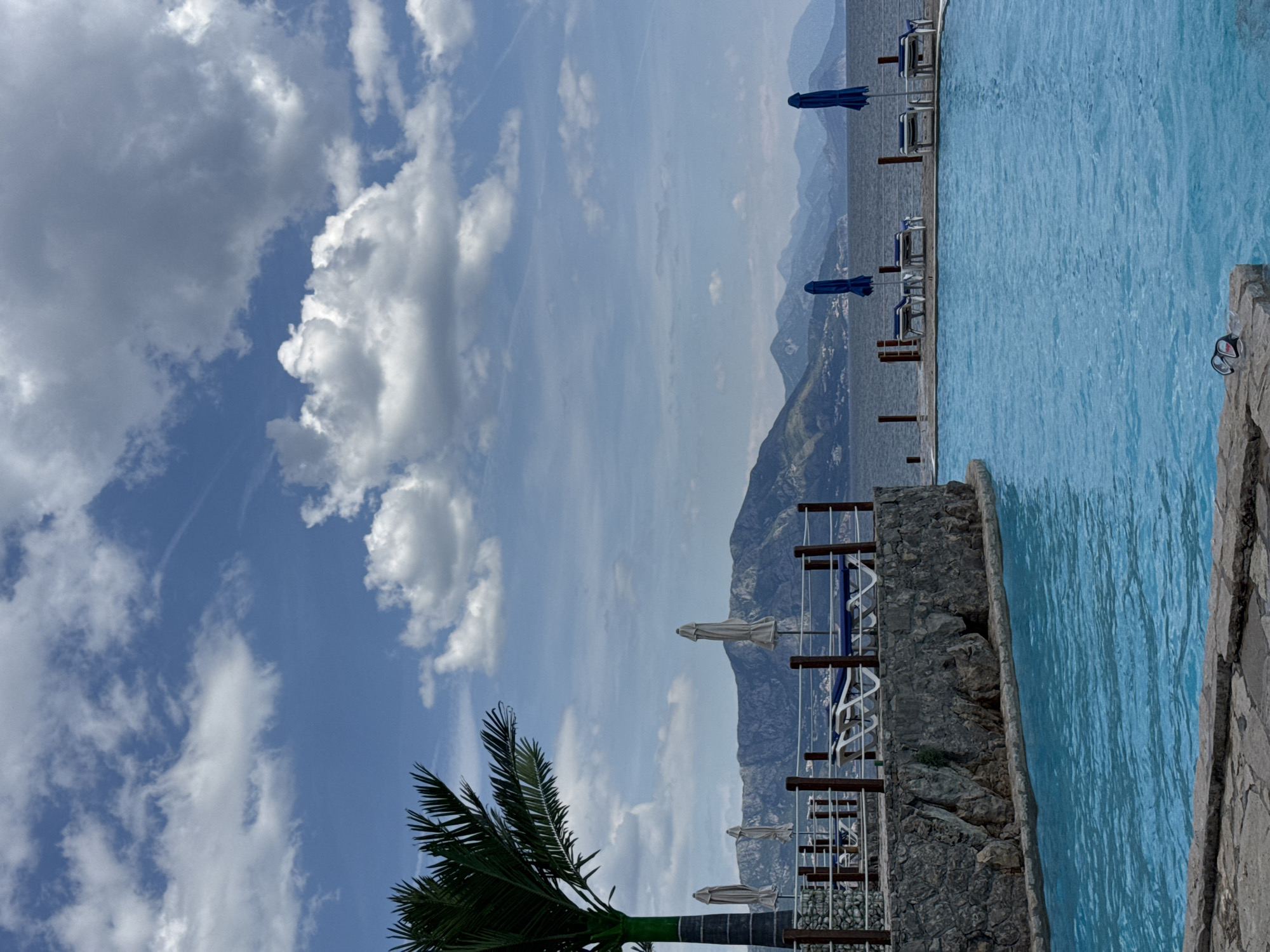 The image shows a scene in Prijevor, Montenegro. In the foreground, there is a swimming pool filled with light blue water. Adjacent to the pool is a stone wall with a railing. Behind the railing, there are lounge chairs with blue umbrellas positioned along the edge. The background features a mountainous coastline, and the sky is partially cloudy with patches of blue. A palm tree is visible on the lower left side of the image.