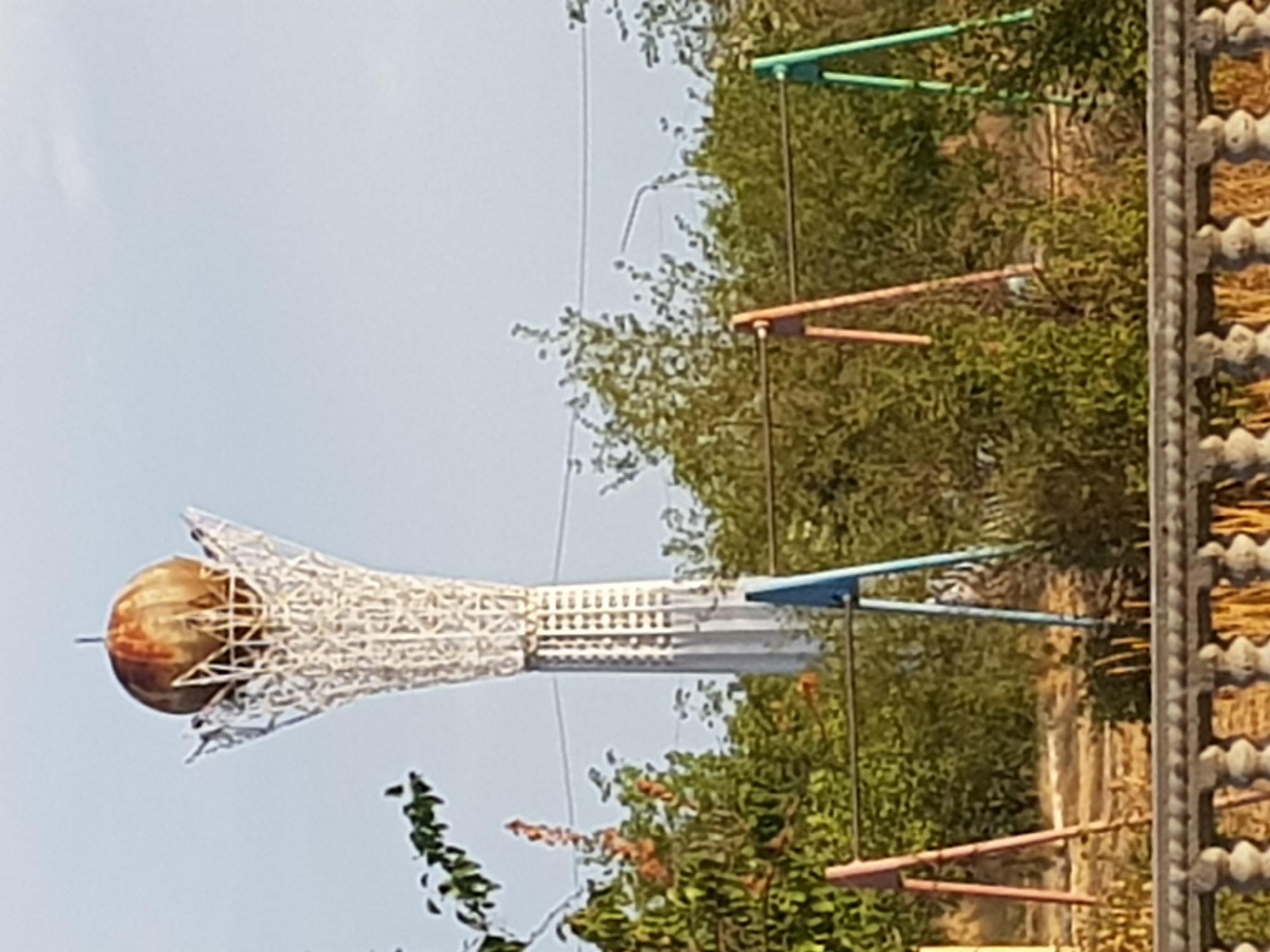 An outdoor, medium-shot image shows a tall, white, lattice-like structure with a rusted, spherical object at its apex, located in the city of Kurchatov, Kazakhstan. The structure, likely a monument or tower, is set against a pale blue sky. In the foreground, a decorative stone railing with balusters separates the viewer from a playground area with colorful metal A-frame structures supporting horizontal bars. Green foliage and trees surround the playground and the base of the tower. The overall scene suggests a public space in a moderately vegetated urban environment.