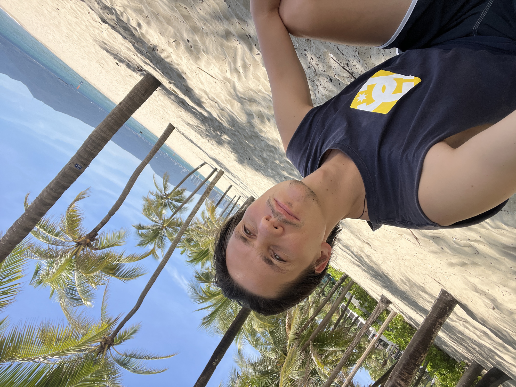 A man with dark hair and a mustache sits on a sandy beach in Đà Nẵng, Vietnam. He is wearing a dark blue sleeveless shirt with a yellow and white logo on the left side of his chest, and black shorts. He is looking towards the camera. Behind him, a row of palm trees lines the sandy beach, with the ocean and mountains visible in the distance. The sky is blue and clear.