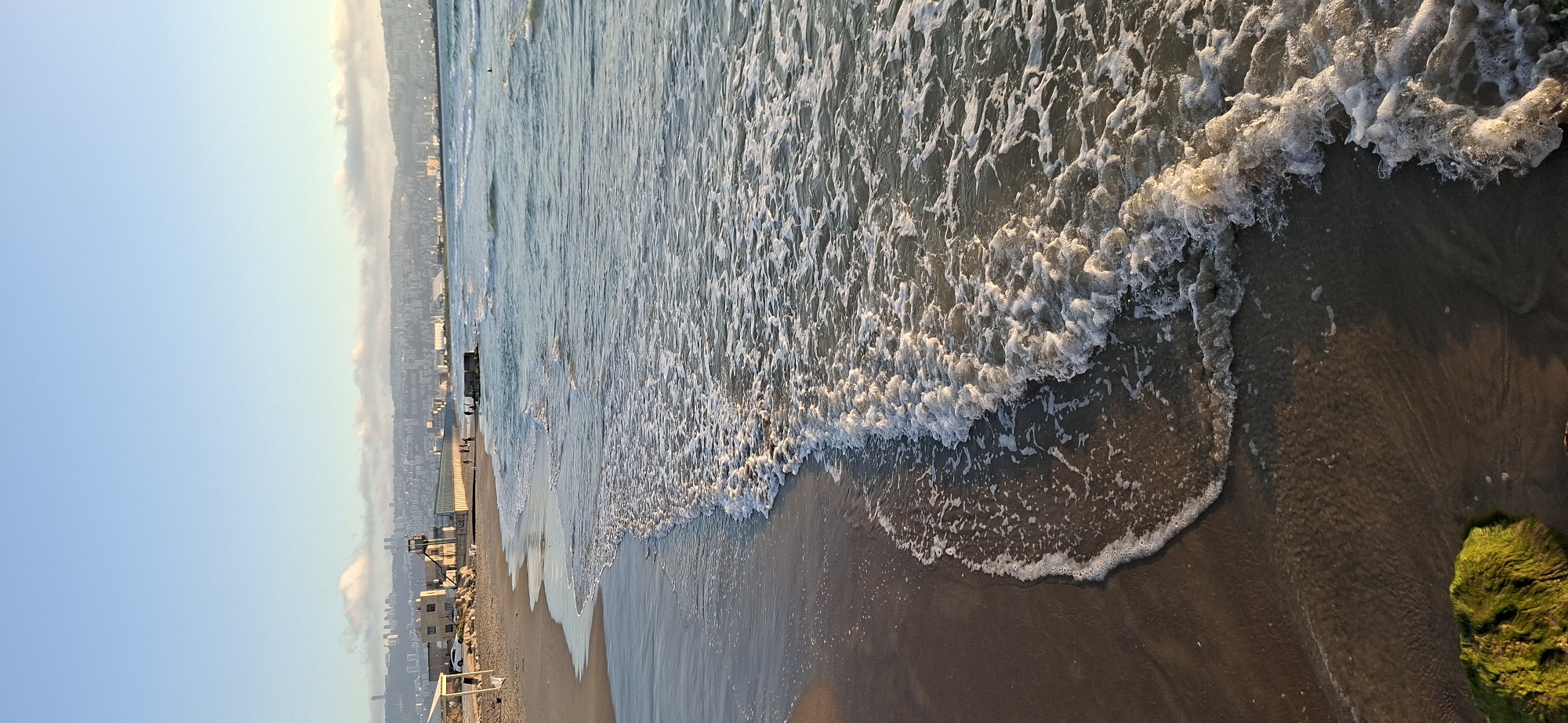 This image depicts a tranquil beach scene with a city skyline in the background. The foreground is dominated by the ocean, with gentle waves lapping at the sandy shore. The water is a pale blue-green, with white foam cresting the waves. The sand is a light brown, wet from the receding tide, and shows ripples from the water's movement.
In the midground, a bustling city skyline stretches along the coast, its buildings appearing muted and slightly hazy, suggesting distance or perhaps a light fog. Several structures, possibly part of a boardwalk or pier, are visible along the shoreline, with a few small figures scattered on them, suggesting human presence and activity. The sky above is a clear, pale blue, fading to a lighter hue near the horizon, indicative of early morning or late afternoon light.
A notable detail is a patch of vibrant green, mossy material on the sand in the bottom right corner of the frame, contrasting with the earthy tones of the beach and water. The overall mood is serene and peaceful, capturing the quiet beauty of a coastal urban landscape. No visible text is present in the image.