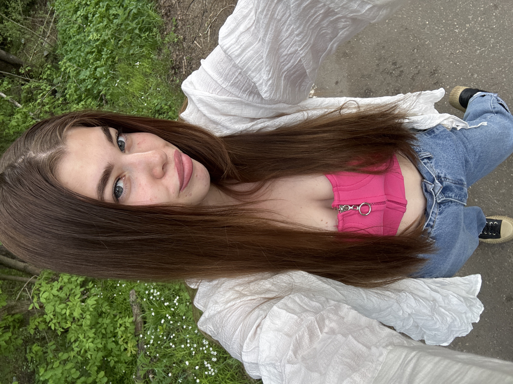 A woman with long brown hair is posing for a selfie outdoors. She is wearing a pink top, blue jeans, a white shirt, and black and beige sneakers. The background consists of green foliage and a concrete surface, possibly a sidewalk or path. The location is unspecified beyond the city of Мещёры, Russia.