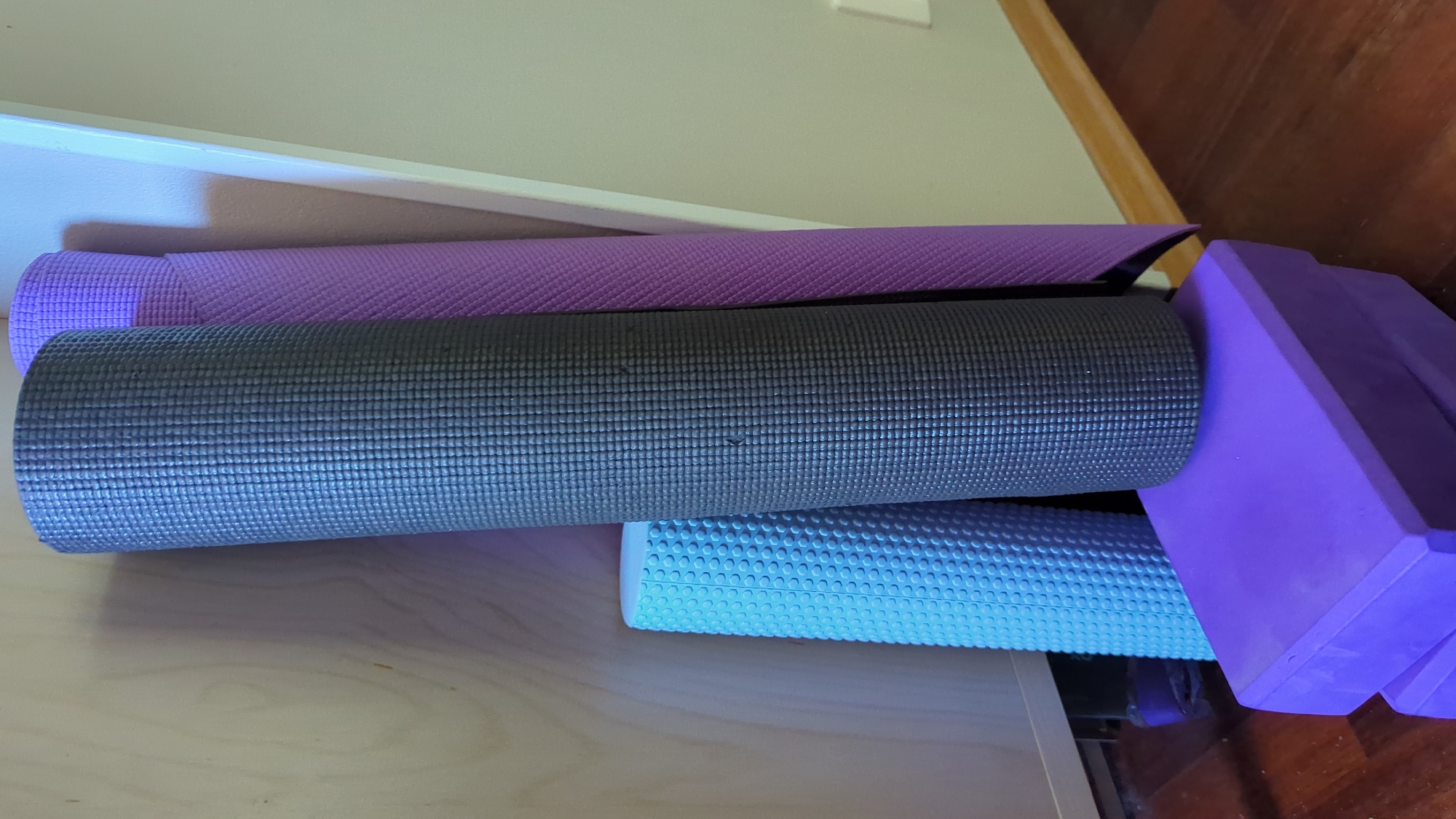 In Kløfta, Norway, a collection of yoga and fitness equipment is neatly arranged. The scene features a dark gray yoga mat rolled and standing upright, positioned in front of a lighter purple yoga mat that is also rolled and standing. To the left, a light blue foam roller with a textured surface is leaning against the gray yoga mat. Below these items, two stacked purple yoga blocks provide a base. The background consists of a light wood-paneled wall and a neutral-colored wall, with a wooden floor visible at the bottom. The overall impression is one of organized athletic gear.