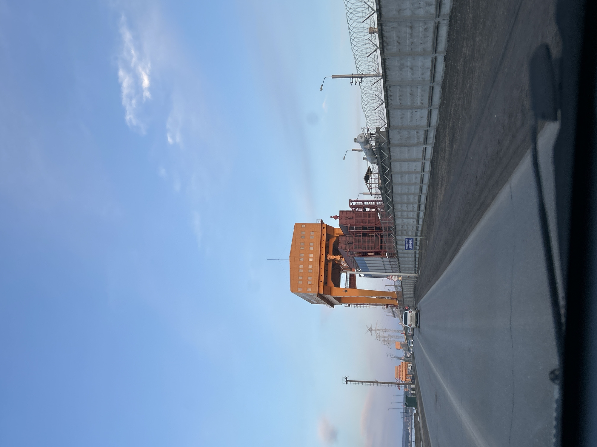 The image shows a view from inside a vehicle, possibly a car or truck, on a road in what is presumably the city of Chaikovsky, Russia, judging by the user's prompt. The sky is blue with sparse, scattered clouds. Alongside the road is a high concrete wall topped with barbed wire. An orange gantry crane is prominent in the middle ground of the scene. Industrial structures are also visible behind the crane. Further down the road, there are power lines and possibly more cranes or industrial equipment. A white vehicle is visible on the road.