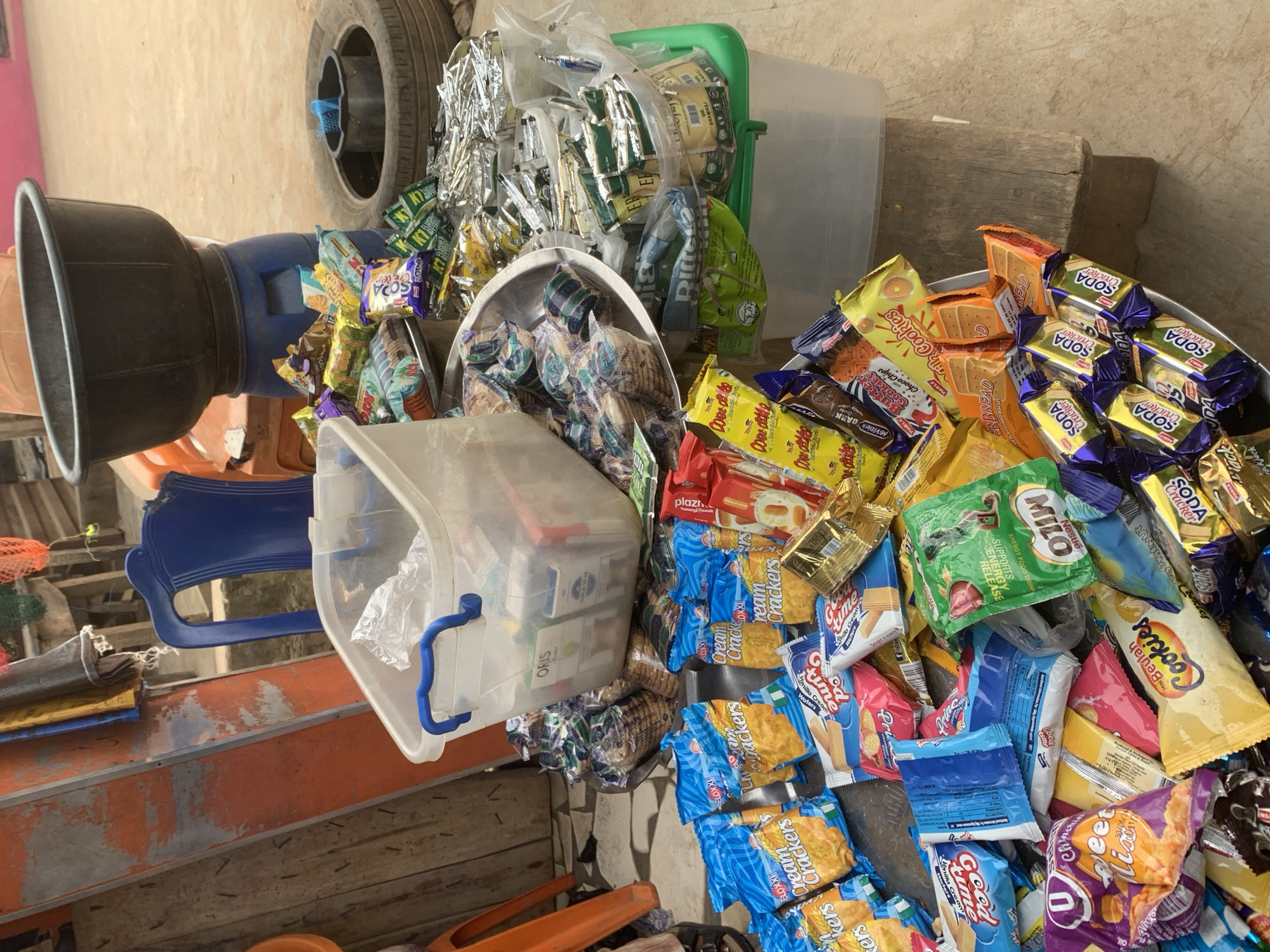 This is a forensic, matter-of-fact description of a street stall in Shagamu, Nigeria, featuring an assortment of packaged snacks and drinks. The stall appears to be situated outdoors, possibly on a dusty or unpaved surface, with a wooden plank serving as a makeshift table for displaying the merchandise. In the background, a blue plastic chair is visible, along with what appears to be parts of a wooden structure, possibly a makeshift stall or building.
The primary focus of the image is a variety of packaged goods, including crackers, cookies, and beverages, arranged in piles and containers. Notably, several packages of "Cream Crackers" are prominently displayed, along with other items labeled "Good Time," "Beylah Cookies," and "Soda Cracker." A large clear plastic tub, filled with more packaged items, sits on top of a metallic serving tray. Next to this, a green plastic container holds additional packaged goods, some of which appear to be beverages. A large black basin rests on top of a blue plastic drum in the background, and a used tire is also part of the scene, possibly repurposed for display or as a container. The overall impression is one of a busy street market stall, typical of many urban environments in Nigeria, where goods are sold directly to consumers. There are no people visible in the frame.
