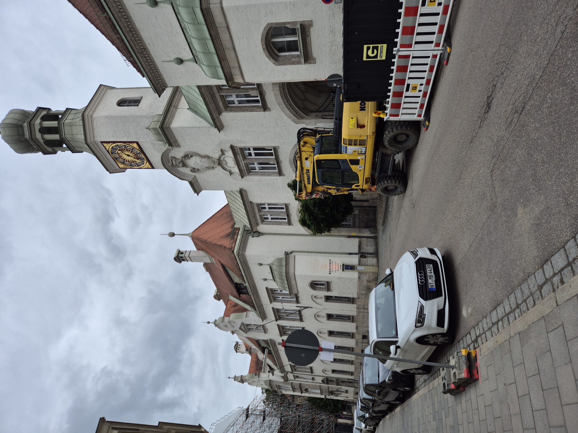 The image depicts a street scene in Regensburg, Germany, with a white building featuring a prominent clock tower. The building is white with a red-tiled roof and has several windows. A large yellow excavator is parked on the street in front of the building, next to a black construction container with a logo that appears to be "GUGGENBERGER". A temporary barrier made of white and red striped panels is also present. Several cars are parked along the sidewalk on the left side of the image, including a white Audi. The sky is overcast, suggesting a cloudy day. A sign on the building reads "BRAUHAUS AM SCHLOSS" which indicates the location.