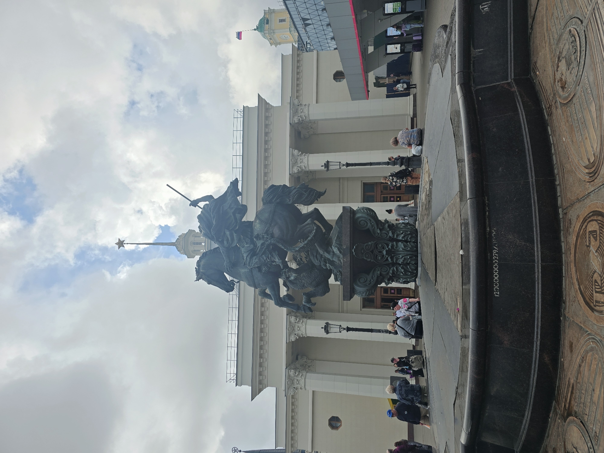 The image shows a monument of a person on horseback spearing a dragon, located in front of a large, white neoclassical building with columns. A Russian flag flies from a tower in the background. The sky is cloudy. Several people are visible in the foreground, some standing and some sitting. The monument appears to be a fountain, with a dark, tiered base. The building in the background is likely a Moscow Metro station, given the architectural style and the star on the spire, which is a common feature of Stalinist architecture in Moscow. Specifically, this is likely the monument to Yury Dolgorukiy in front of the Rossiya Cinema (now the Zaryadye Hall of Music) and also near the Rossiya Hotel (demolished). The fountain itself is actually the "Friendship of Peoples" fountain, but it's not the main one, this one is a replica of it. The original "Friendship of Peoples" fountain is located in VDNKh park. The monument in the image is the monument to Yury Dolgorukiy, founder of Moscow.