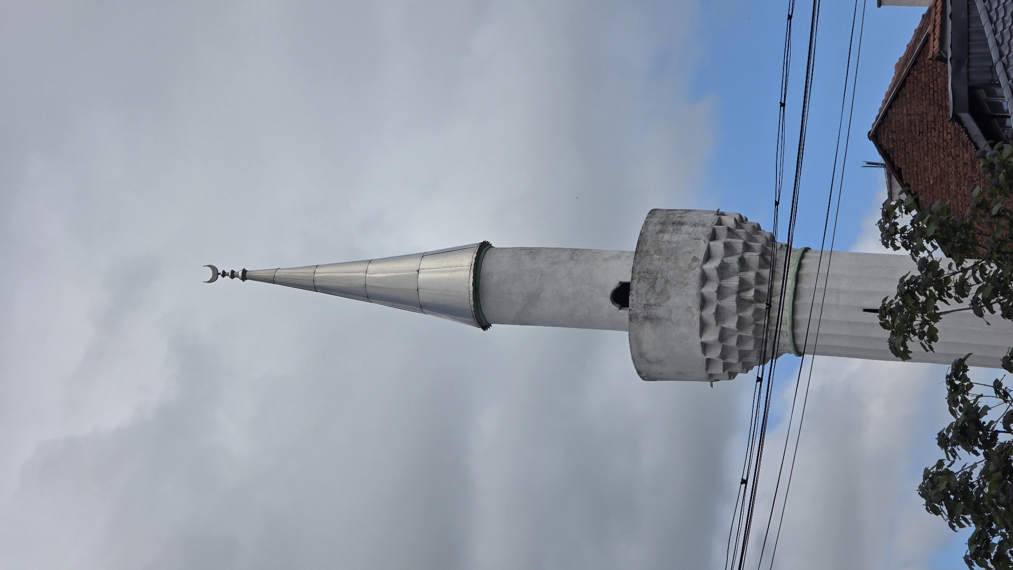 A vertical shot shows the minaret of the Orta Dzhamiya mosque, situated in Blagoevgrad, Bulgaria. The minaret is light grey, with a metal conical roof topped by a crescent moon. It features a textured ring around the top section. Electrical wires run across the top right of the frame, and parts of a brick building and green foliage are visible on the right side. The sky, a mix of blue and grey, forms the background.