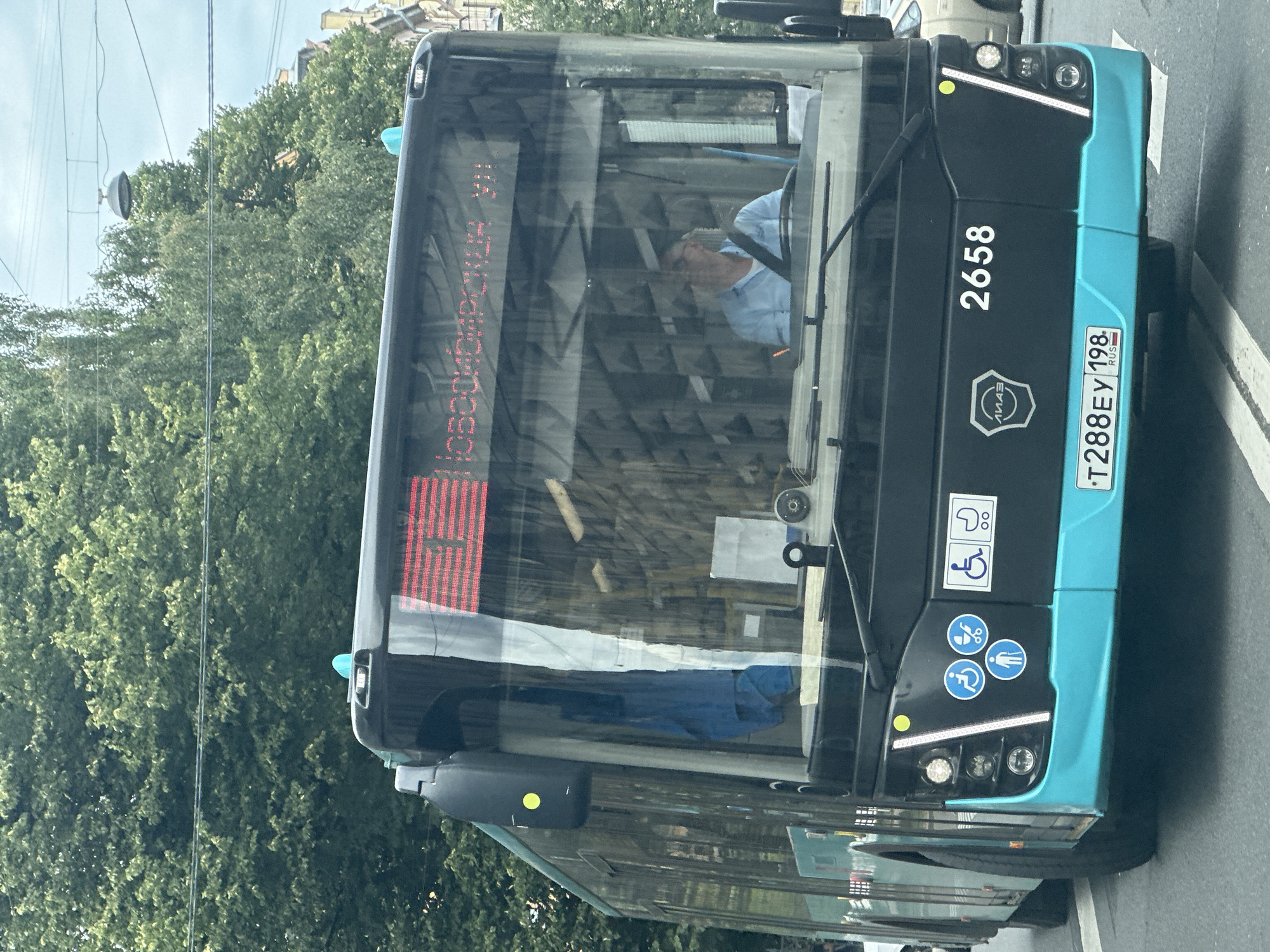 An eye-level, slightly low-angle, medium shot shows the front of a teal and black public bus driving on a street in Saint Petersburg, Russia. The bus, a PAZ-4234 model, has a digital display on its windshield showing red lettering in Cyrillic that reads "Новосибирьская VII" (Novosibirskaya VII). A male bus driver wearing a light blue collared shirt is visible behind the windshield, looking to the right. Below the windshield are several blue and white circular stickers: one depicting a person in a wheelchair, another showing a stroller, and a third showing a person walking. To the right of these stickers are two white rectangular stickers, one showing a person in a wheelchair and the other a stroller. The bus displays the number "2658" in white digits on the black section of its front. The license plate of the bus reads "T288EY 198 RUS". The bus is surrounded by lush green trees and utility wires are visible overhead, suggesting an urban environment. The bus is driving on a paved road with white lane markings. The overall lighting is overcast, indicating a cloudy day.