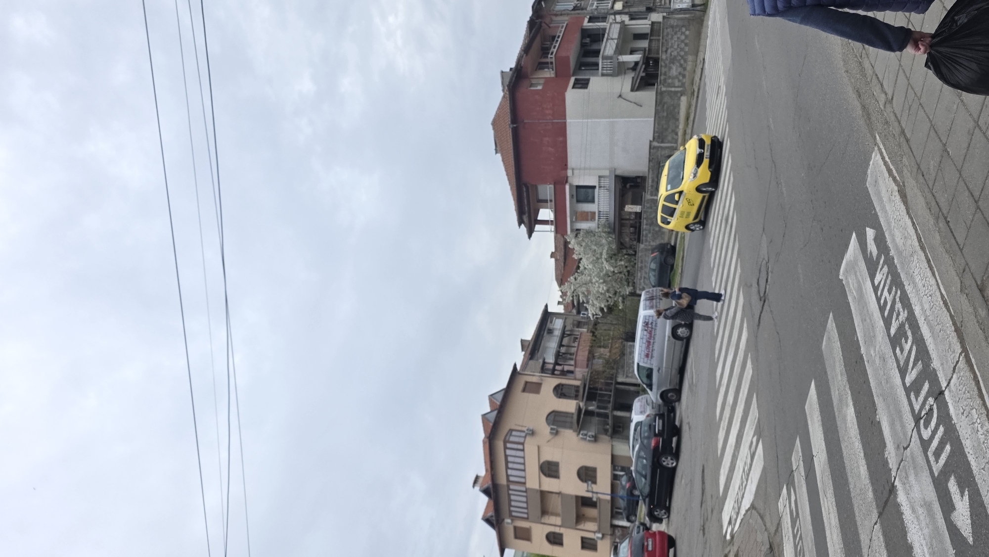 The image depicts a street scene in Blagoevgrad, Bulgaria. The view is oriented along a crosswalk with pedestrian markings written in Bulgarian. A person is crossing the street. Various vehicles, including a yellow taxi and a white van, are parked or moving. Buildings line the street. Overhead, there are several wires or cables. The sky is overcast.