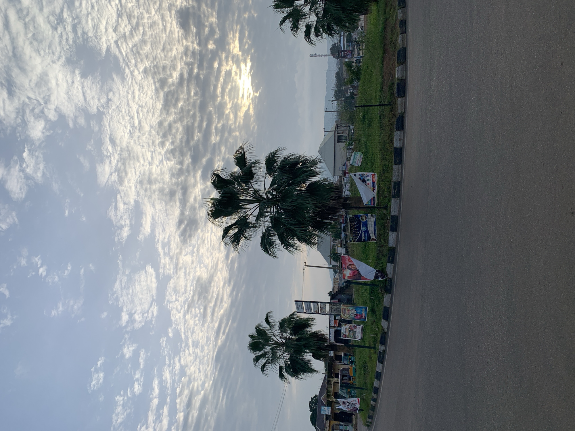 The image captures a street scene at a roundabout in Rafin Par, Nigeria, under a cloudy sky with the sun peeking through. In the foreground, a paved road curves around a grassy central island. The island is adorned with several palm trees and a collection of posters, some of which appear to be political campaign materials or advertisements. A black and white striped curb delineates the edge of the roundabout. In the background, various buildings, including what appears to be a residential structure with a white roof, and a telecommunications tower are visible. Several small vehicles, possibly auto-rickshaws, are parked on the side of the road. The overall impression is a typical urban setting in Nigeria, with the sky indicating either early morning or late afternoon.