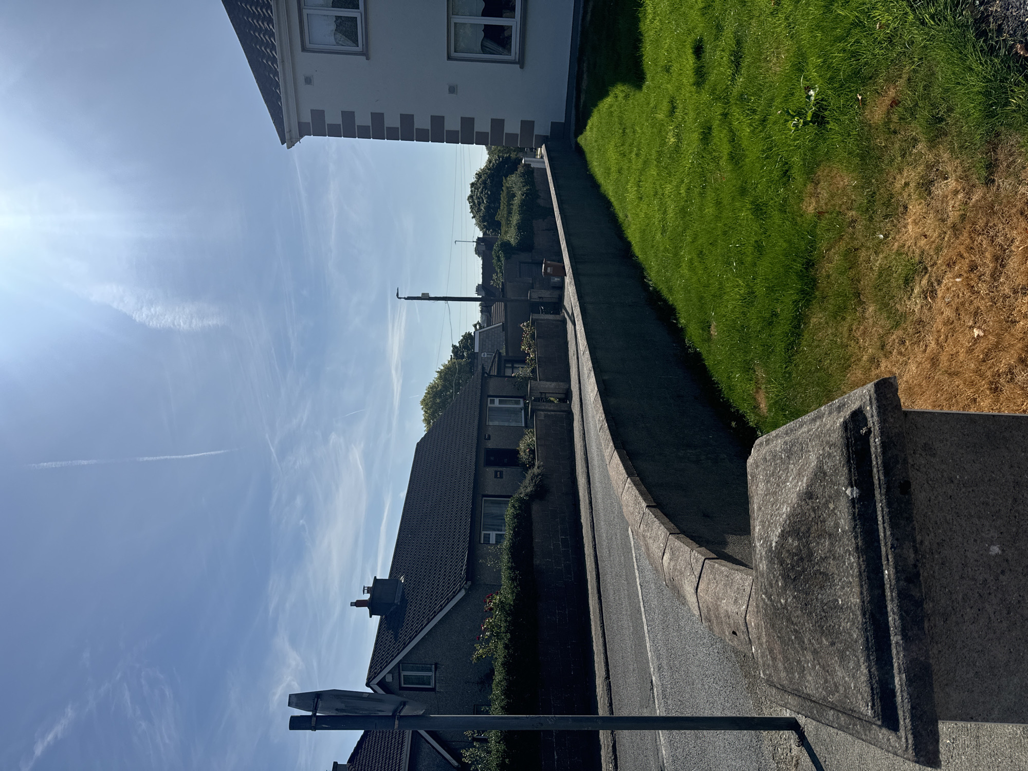 The image depicts a sunlit street scene in Waterford, Ireland. In the foreground, a concrete post with a pyramidal cap marks the edge of a property. To the left, a grey stone wall curves along a paved road. Beyond the wall, a row of houses with dark tiled roofs and grey exteriors line the street. Some houses have well-maintained hedges and gardens. To the right, a modern white house with grey trim is visible. A patch of vibrant green grass slopes upwards from the wall, contrasting with an area of dry, brown grass closer to the foreground. The sky is a clear blue with wispy white clouds and a bright sun, casting strong shadows. A tall metal pole with a light fixture stands further down the street. The overall impression is of a suburban residential area on a bright, clear day.
