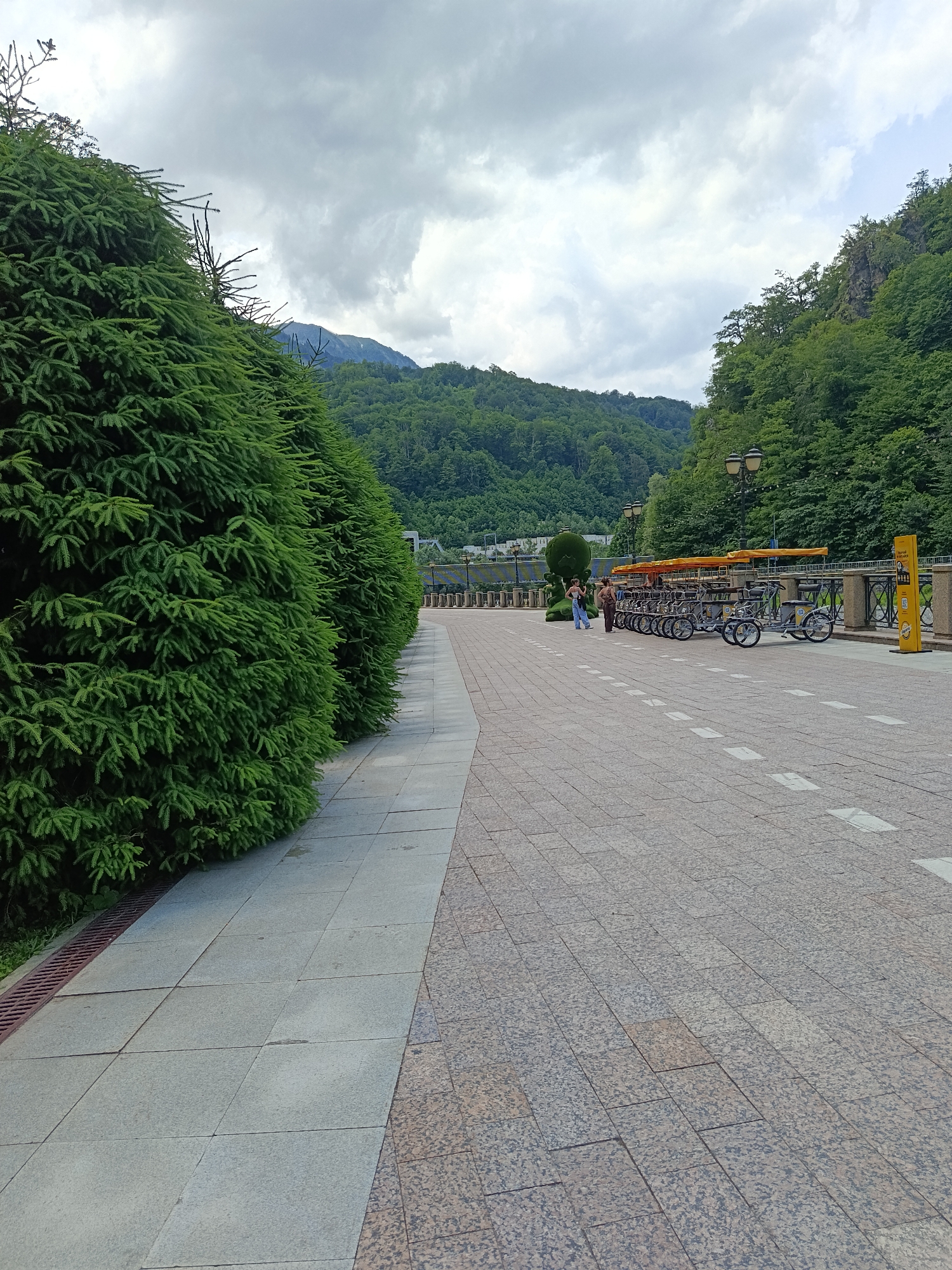 A paved walkway lined with manicured evergreen trees runs diagonally from the left foreground to the center midground of the image. The walkway is composed of two types of paving stones: light grey square tiles on the left and darker, speckled tiles on the right. In the midground, a row of multi-person bicycles, some with orange canopies, are parked along a railing. Two people, a woman and a man, are standing near the bicycles. Beyond the bicycles, a decorative fence separates the walkway from a body of water and the forested slopes of mountains under a cloudy sky. The mountains are densely covered with green trees, and a peak is visible in the distance. The scene appears to be in the resort town of Estosadok, Russia, known for its natural beauty and recreational activities.