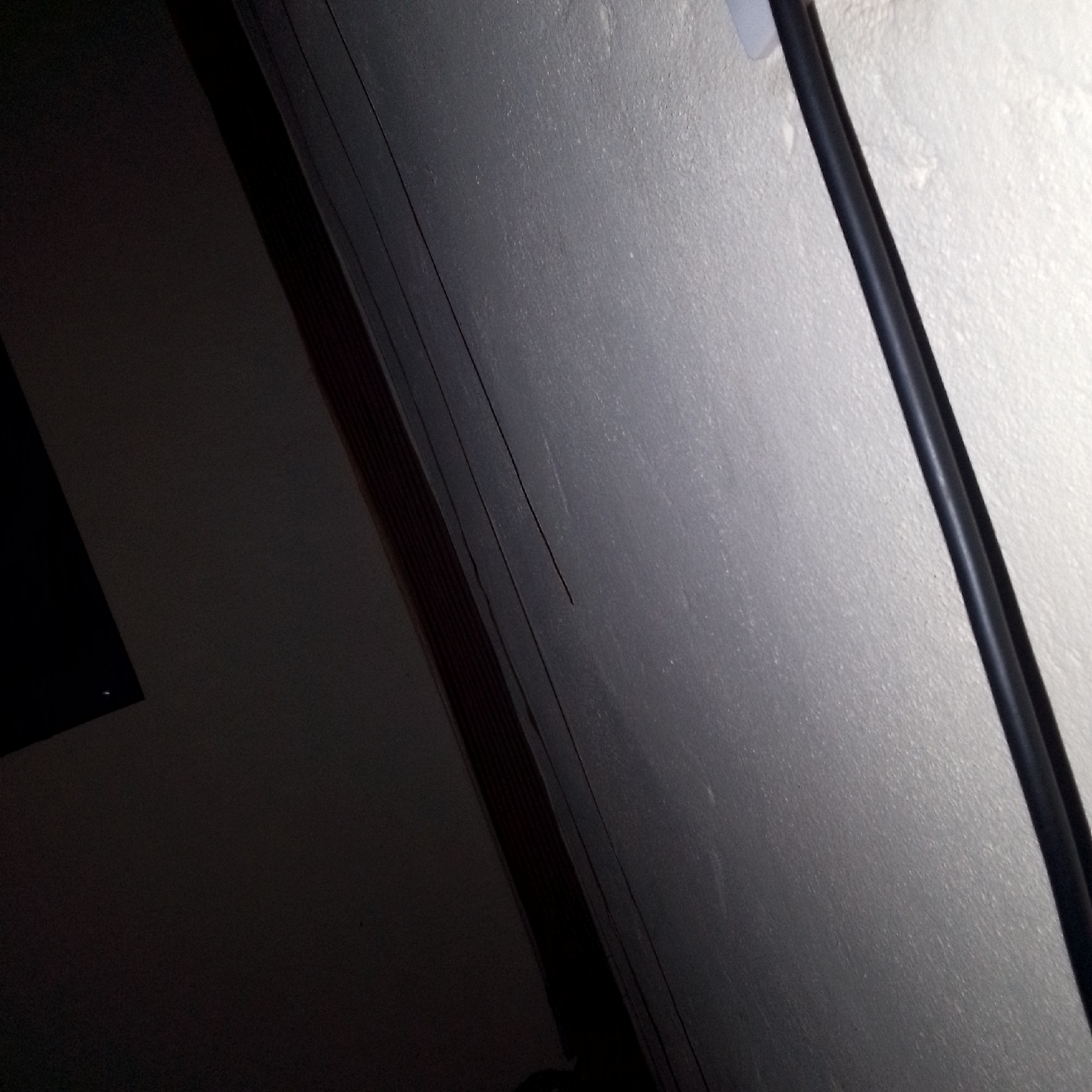 An indoor, angled, close-up shot shows a textured, light gray wall on the right, running diagonally into the frame from the top right corner. A dark, cylindrical object, possibly a cable or pipe, runs parallel to the wall's edge down the right side of the image. On the left side of the image, a dark area obscures much of the scene. A thin, dark line, possibly a crack or seam, is visible on the gray wall. Another dark, rectangular object is partially visible in the upper left corner. The lighting is dim and appears to be artificial. The scene is indoors and appears to be a domestic or commercial interior, though the specific location is unknown.