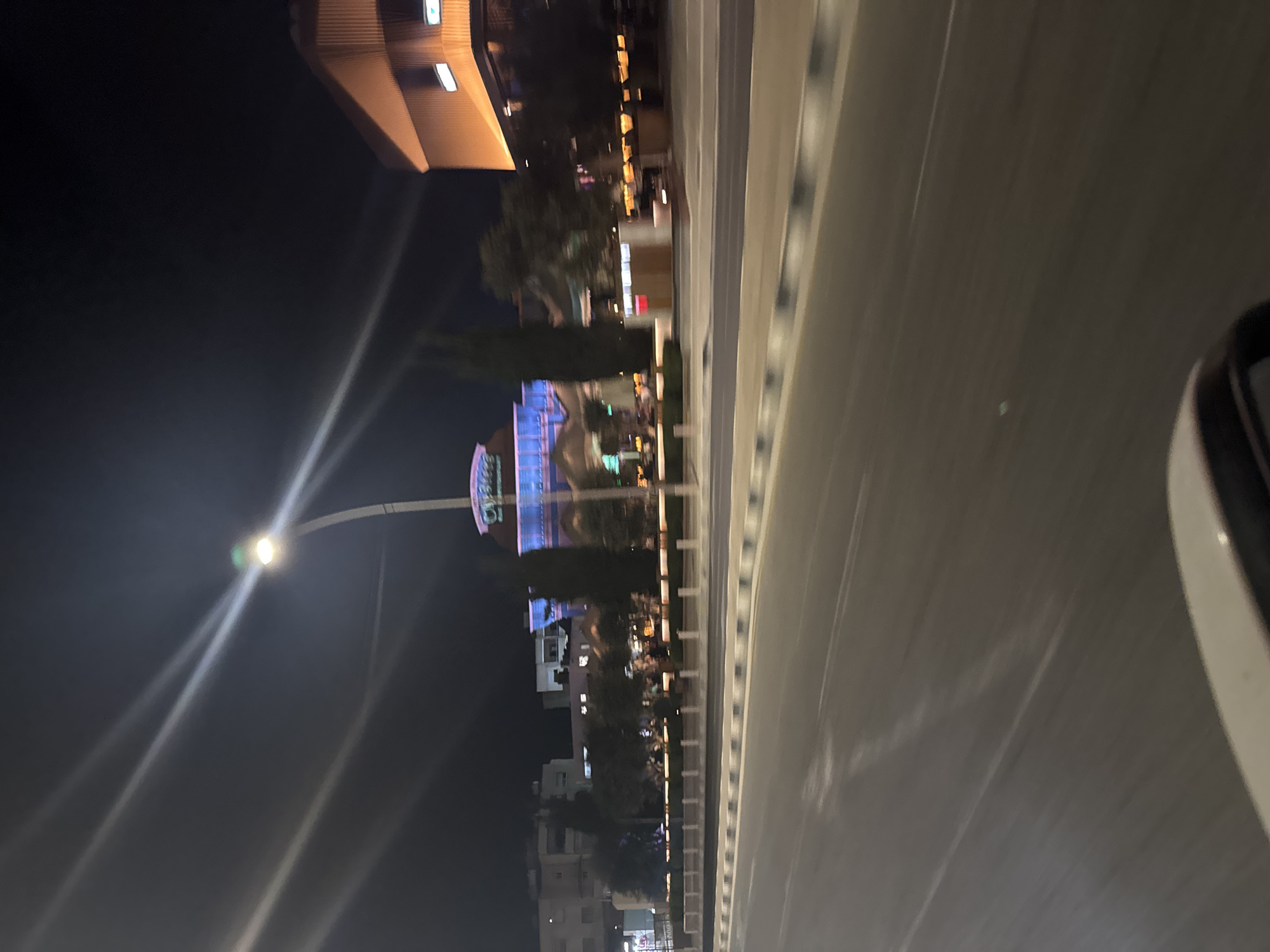 The image captures a nighttime street view in Parekklisia Community, Cyprus. In the background, a building with blue neon lighting and a prominent "Crystal" sign is visible. To the right of this building is a larger structure with warm orange lighting. The street in the foreground is dark, with streetlights casting long streaks of light across the frame due to motion blur. A portion of a vehicle's interior, likely a dashboard or windshield frame, is visible in the bottom right corner. Trees and decorative white bollards line the sidewalk. The overall impression is of a commercial or entertainment area at night, viewed from a moving vehicle.